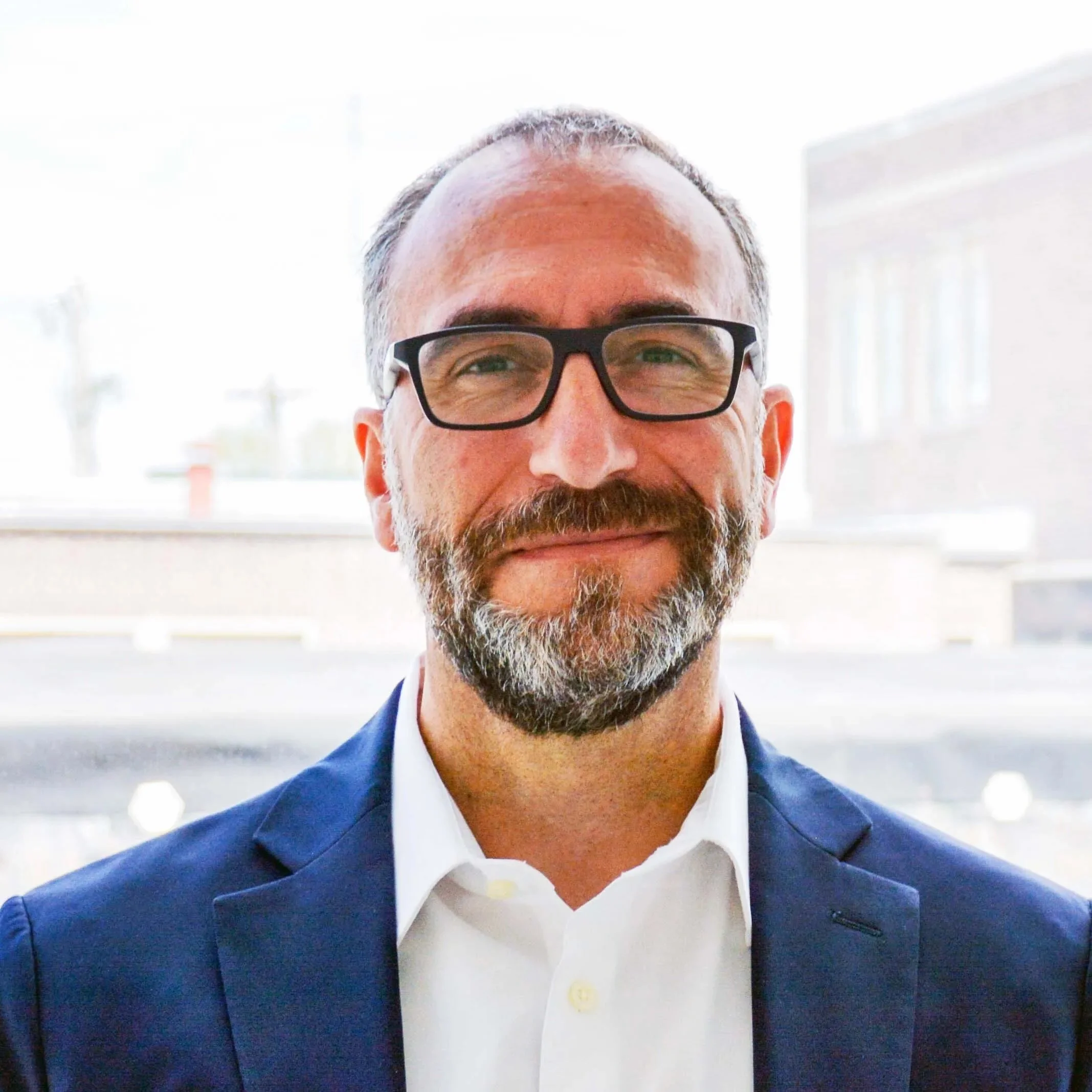 A man with a beard, white shirt, glasses and blue blazer smiling at the camera.