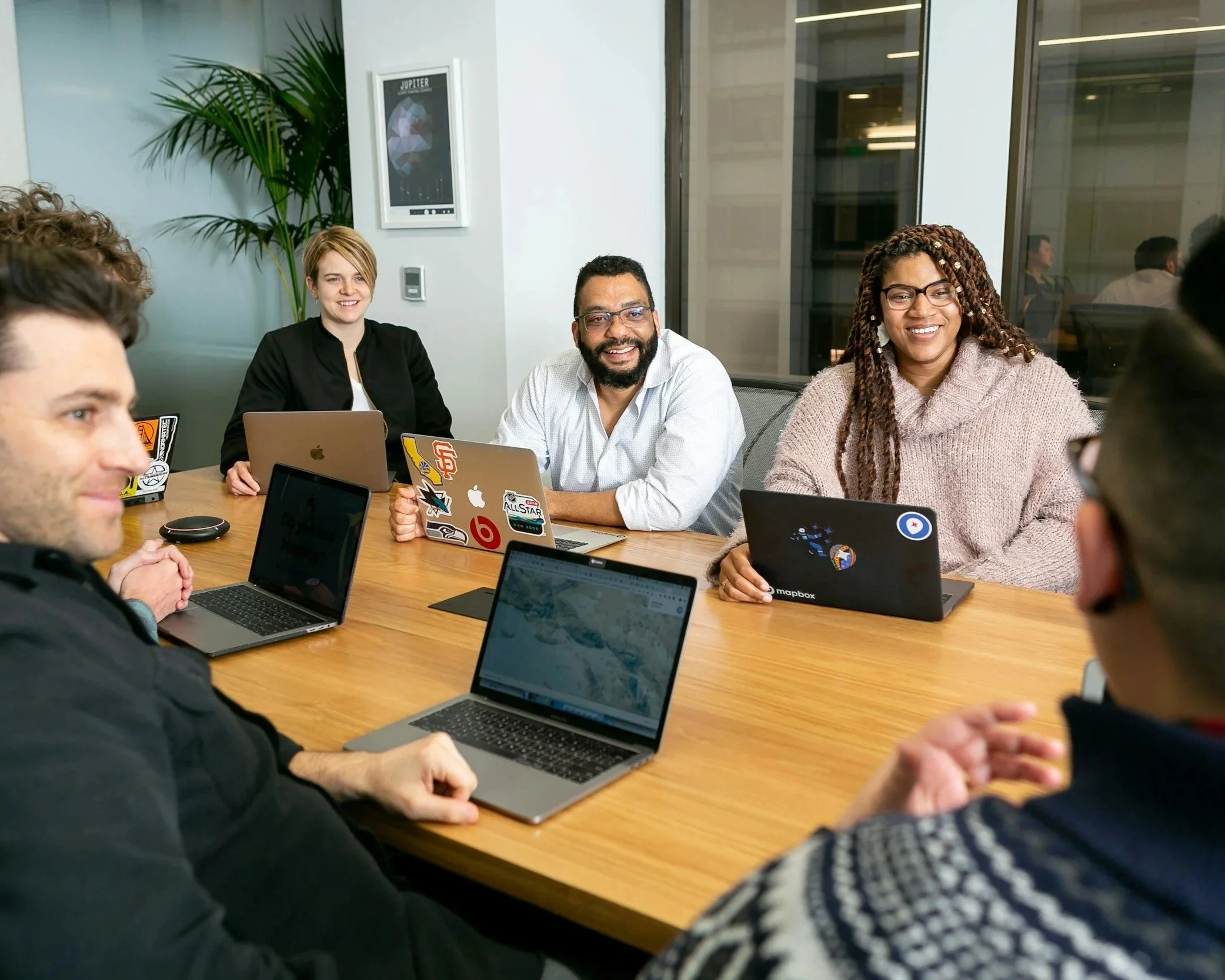 A group of multicultural people sitting around a table with laptops open, likely in a meeting. 