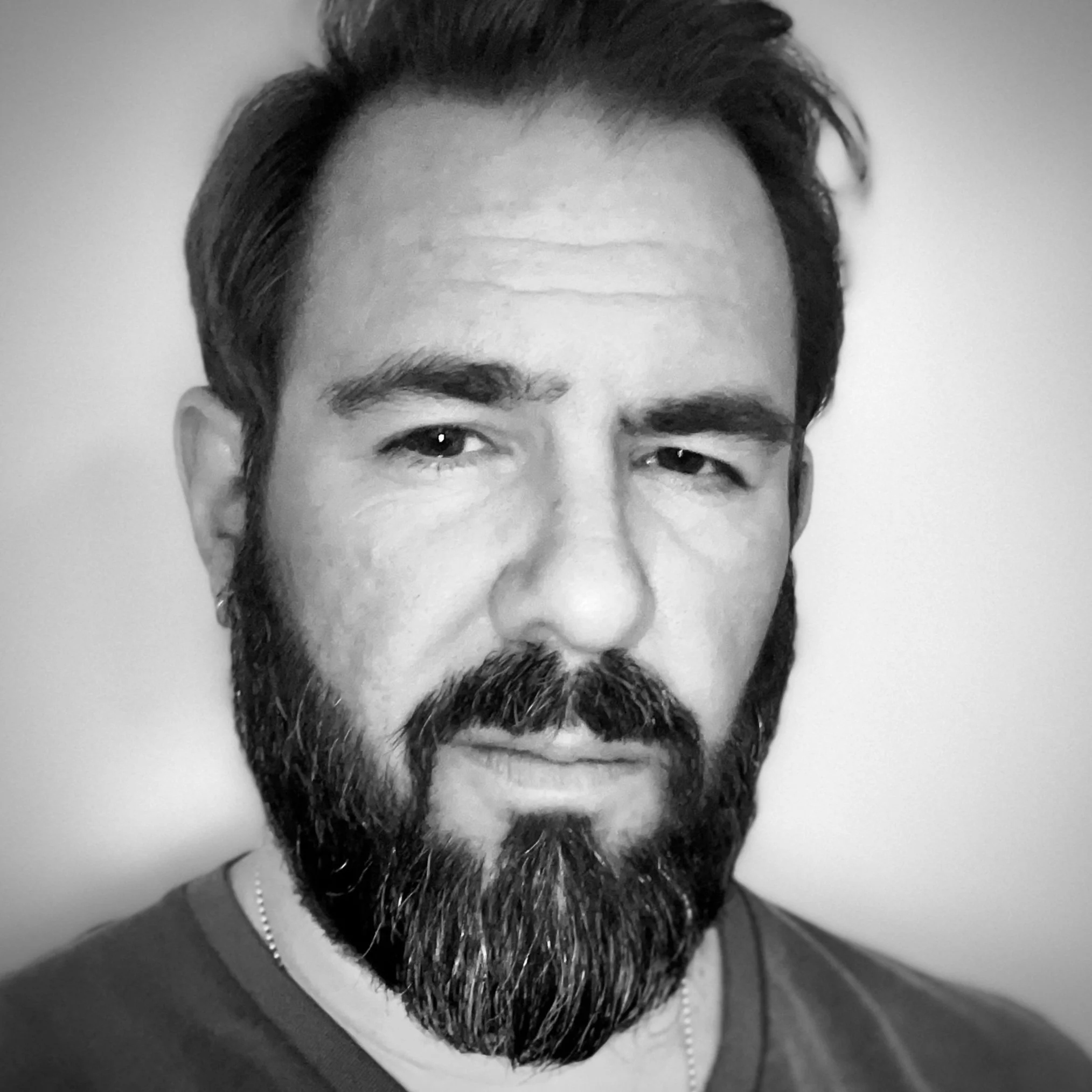 Black and white portrait of a man with a full beard and mustache, wearing a t-shirt and a necklace.