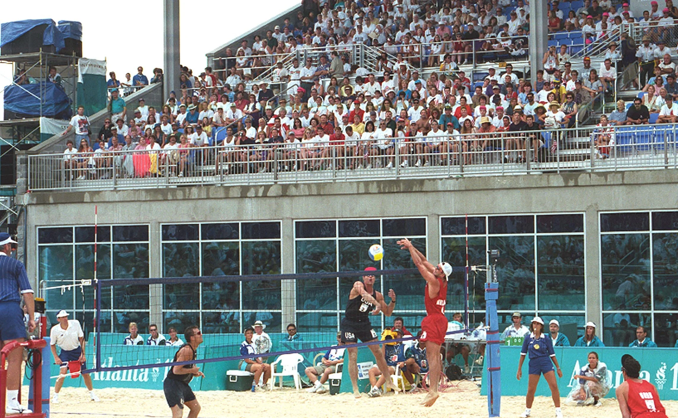 Beach volleyball match with players jumping for the ball, spectators in the stands, officials and coaches around, set on a beach with sand, against a background of a glass-fronted building and grandstand seating.