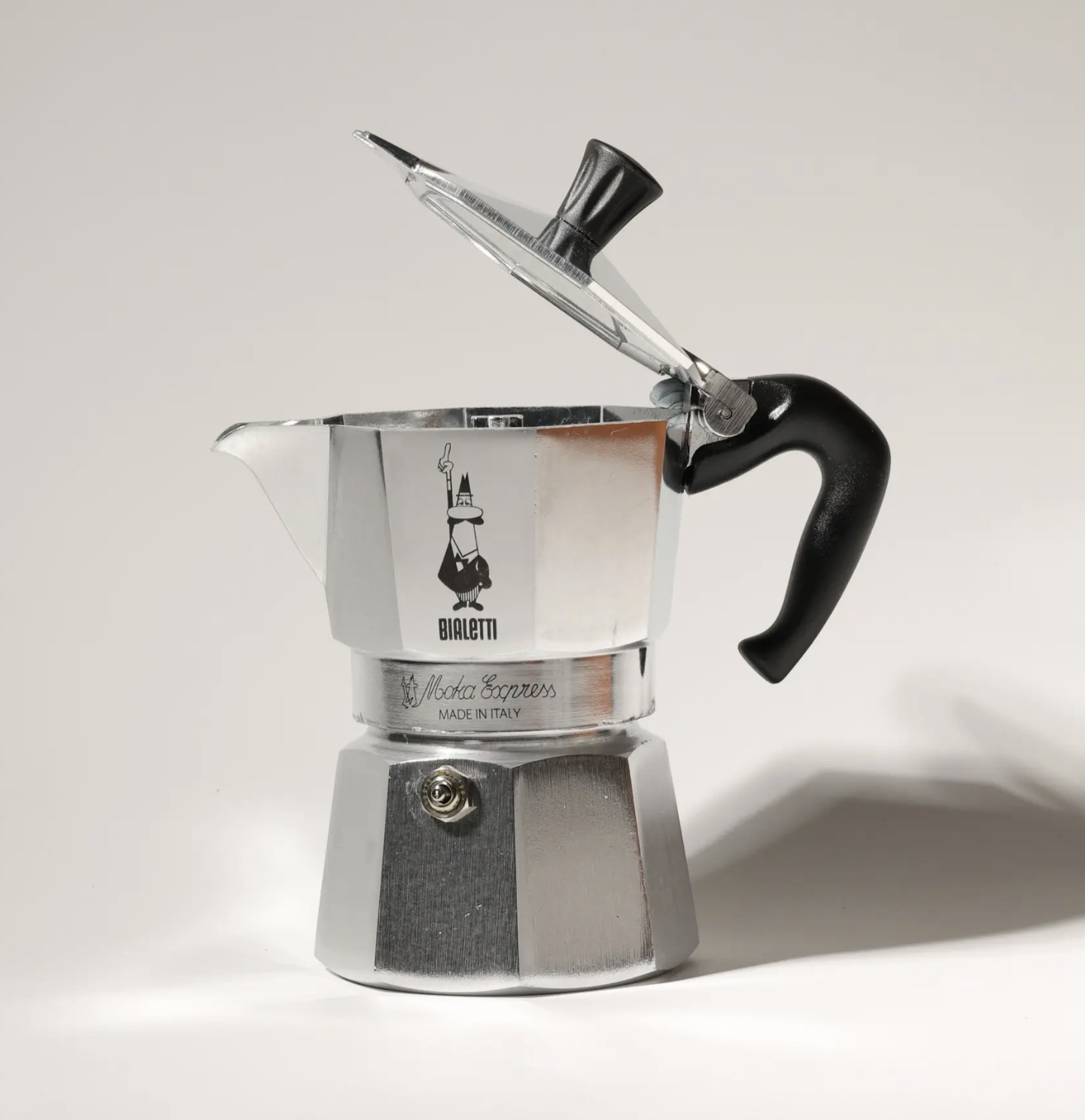 A stainless steel moka espresso coffee maker with a black handle, a hinged lid, and a logo on the front, placed against a plain white background.