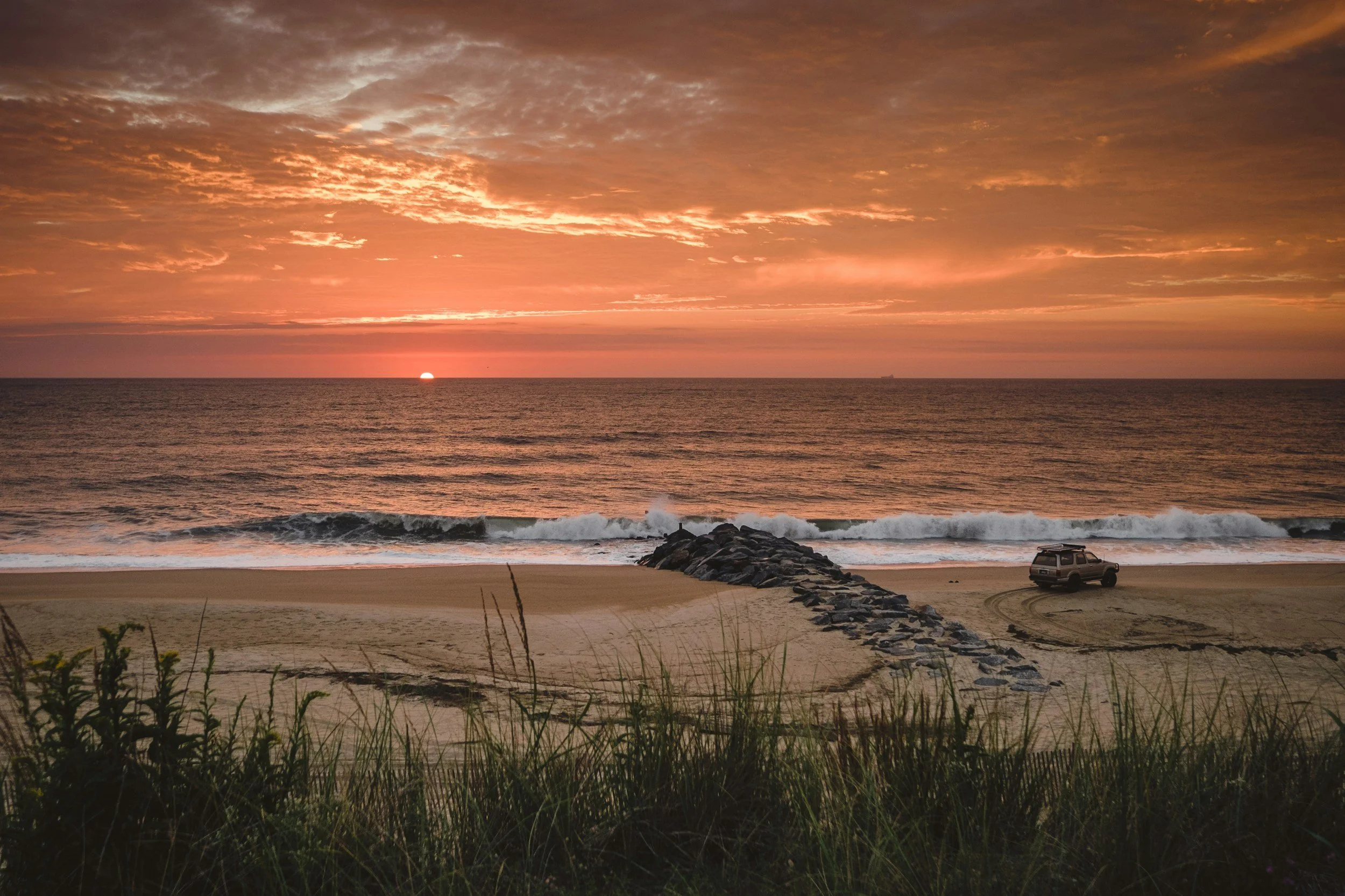 Sunset over ocean with waves crashing on sandy beach, a car parked on the beach near a stone jetty, and grass in the foreground.