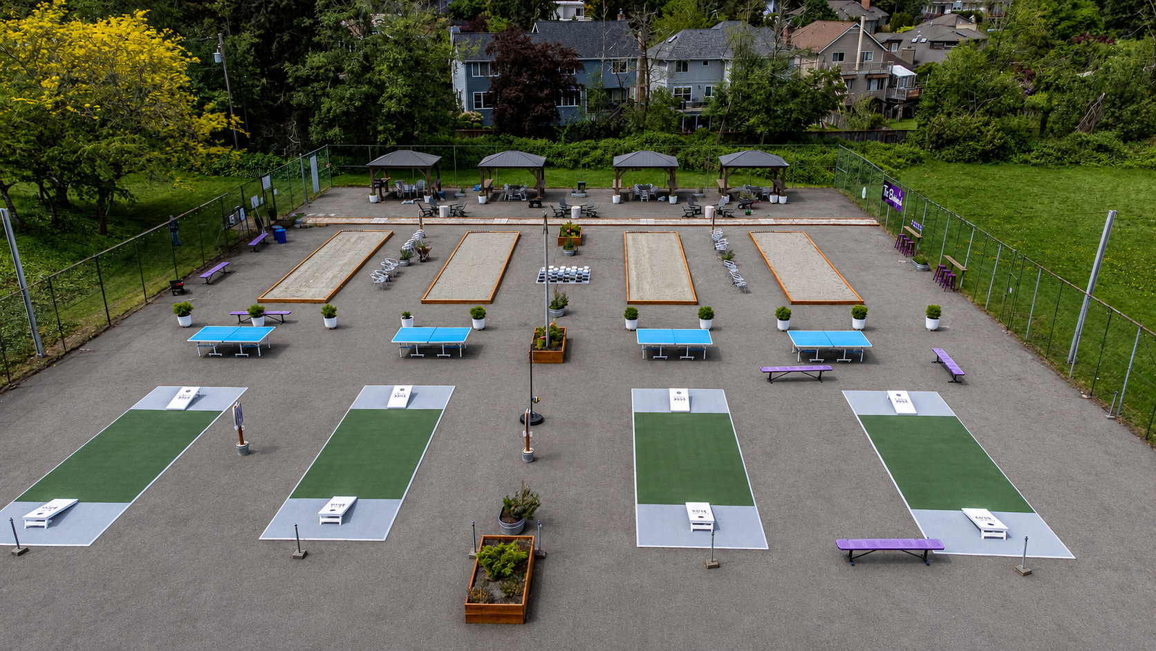 Outdoor park -the Backyard- with cornhole game boards, ping pong tables, benches, potted plants, and shaded seating areas surrounded by a fence and trees.