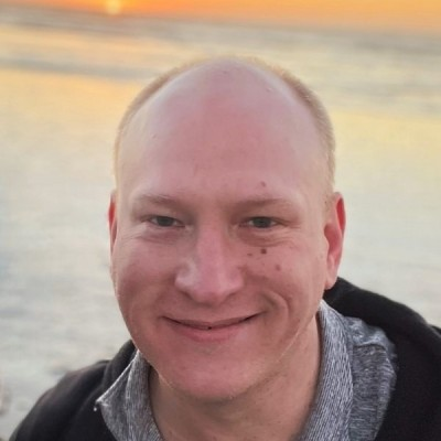 A woman with long brown hair and blue eyes smiling, wearing a dark blazer and earrings, standing in a modern office corridor.