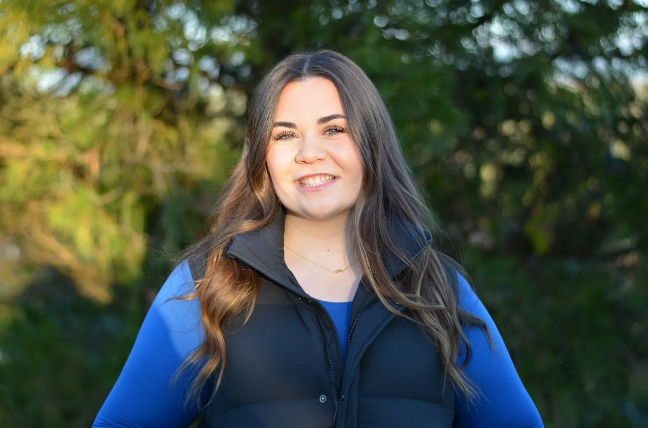 A young woman with long wavy brown hair and light skin, smiling outdoors with trees in the background, wearing a black vest over a blue long sleeve top.