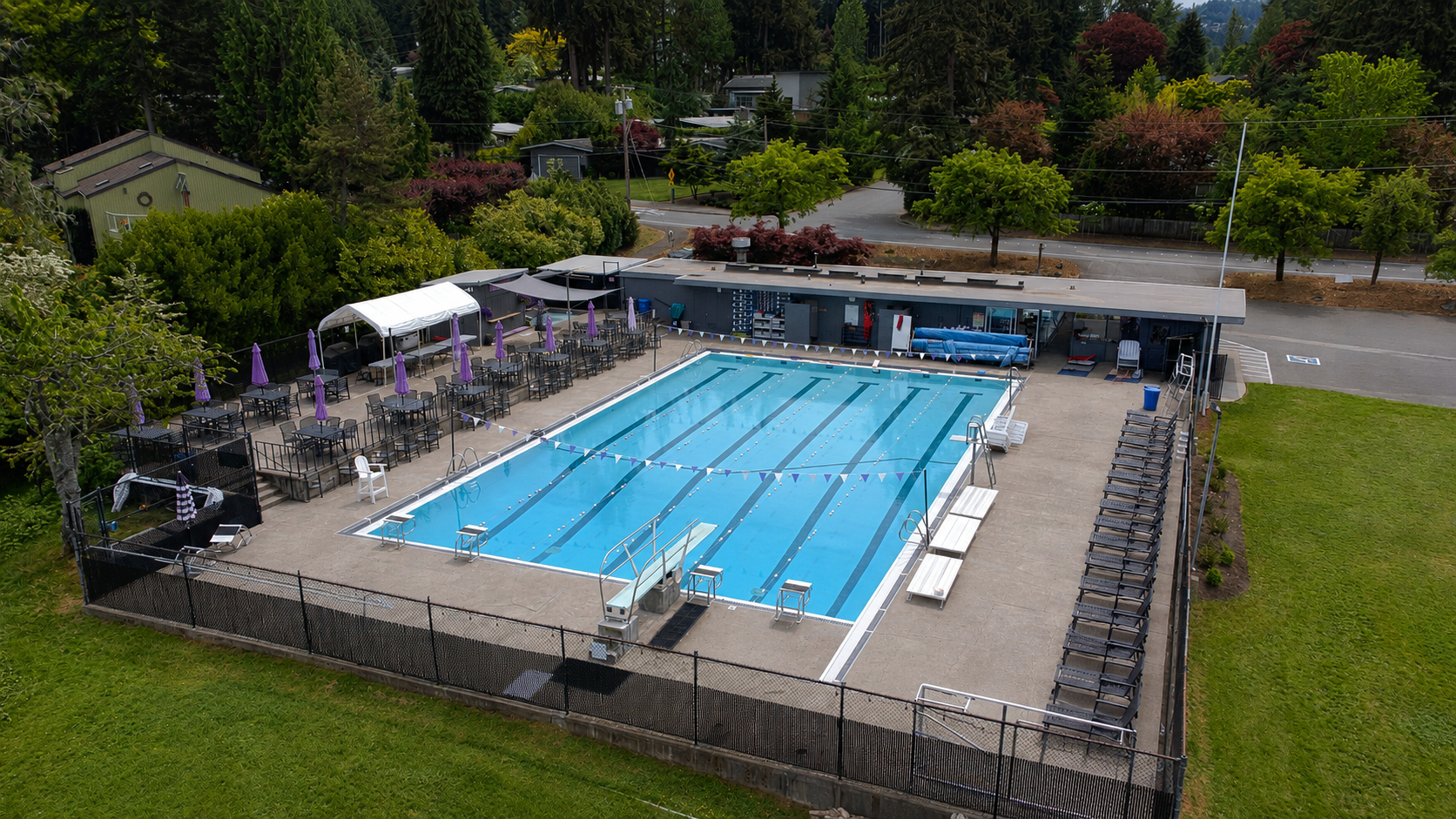Aerial view of the Phantom Lake Club outdoor swimming pool with surrounding lounge chairs, tables with purple umbrellas, and a shaded building. Trees and roads are in the background.