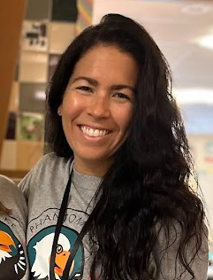 Smiling woman with shoulder-length blonde hair, wearing a navy and white striped long-sleeve shirt, against a plain light background.