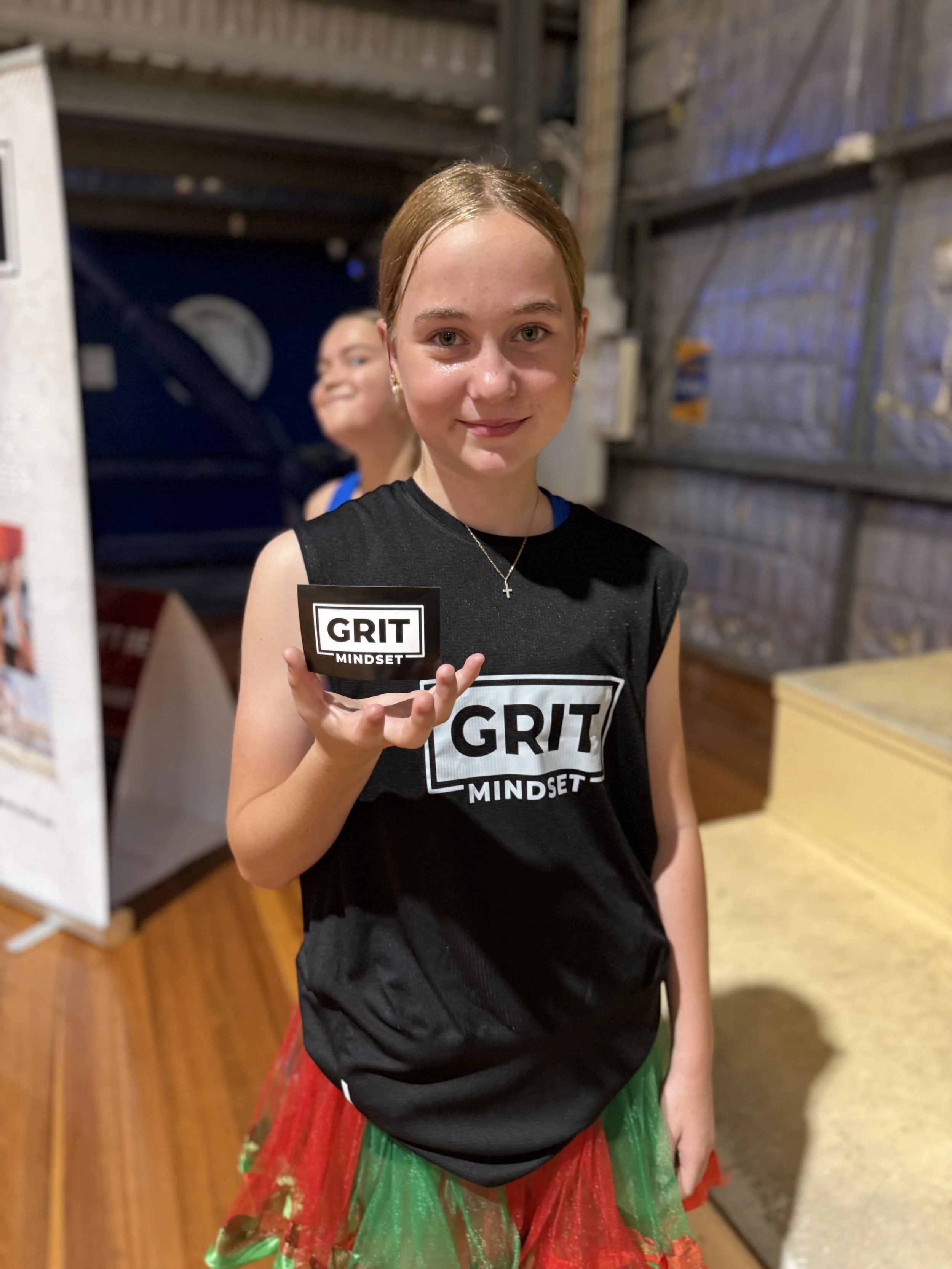 A young girl with reddish hair and fair skin is holding a small sign that says "GRIT MINDSET". She is wearing a black sleeveless shirt with the same message, and a colorful red and green skirt. She is indoors, with a wooden floor and a background tha