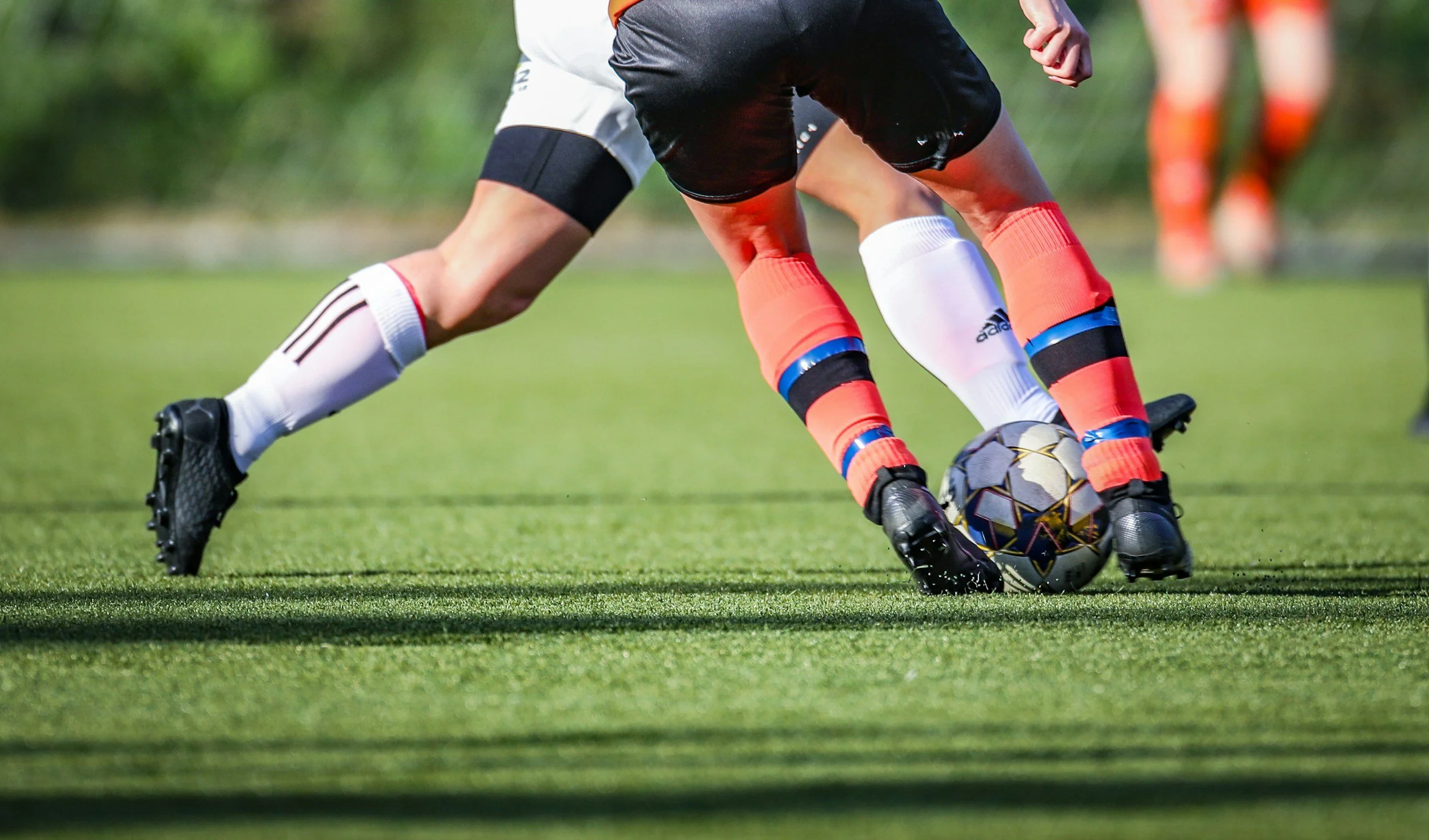 Close-up of two soccer players competing for the ball on a green field, with one player wearing black shorts and white socks, and the other wearing orange socks and black shorts, with a third player in the background.