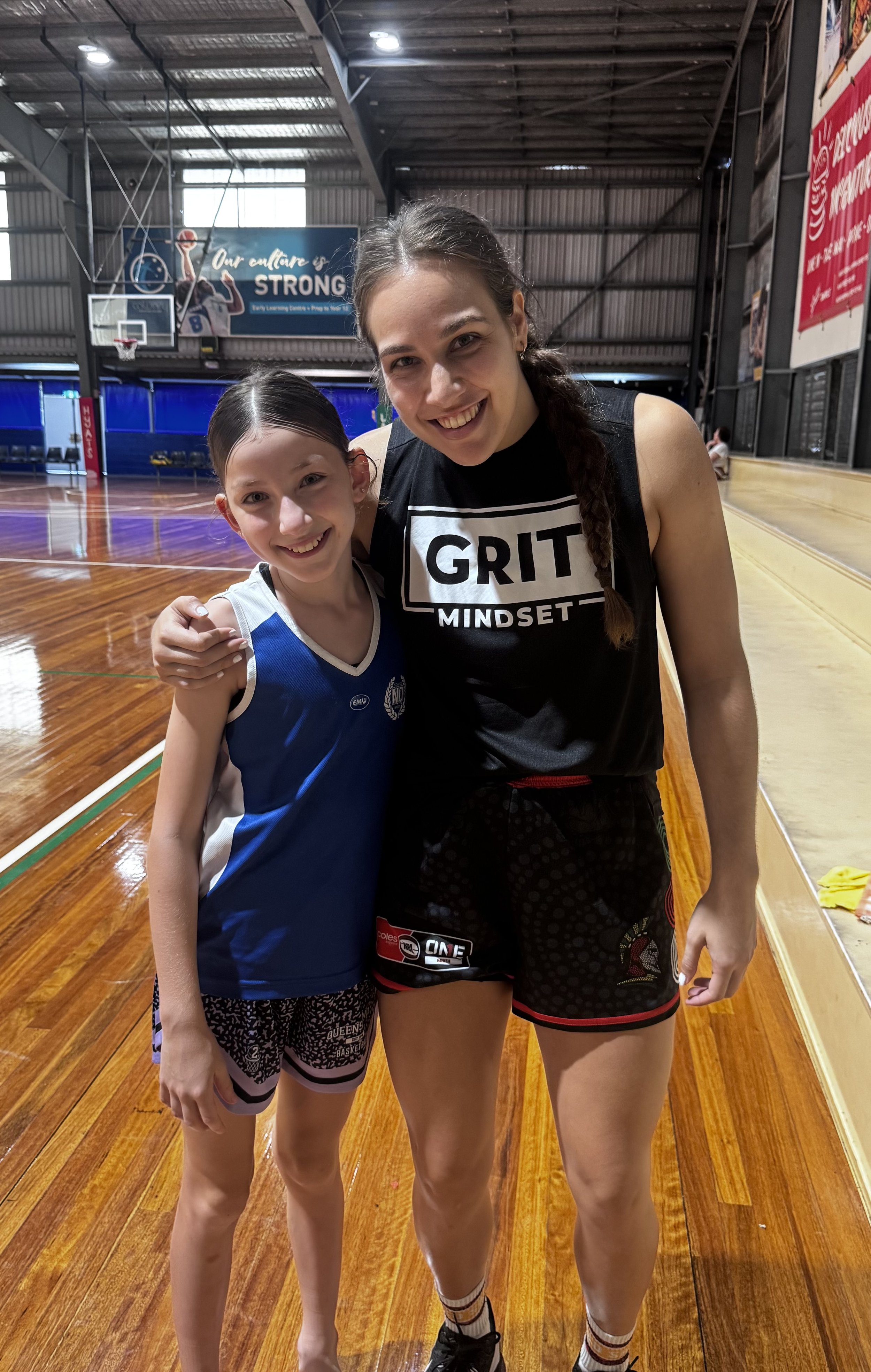 A woman and a girl standing together in an indoor basketball court, smiling at the camera. The woman is wearing a black sleeveless top with 'GRIT MINDSET' written on it and black shorts, while the girl is wearing a blue and white basketball uniform a