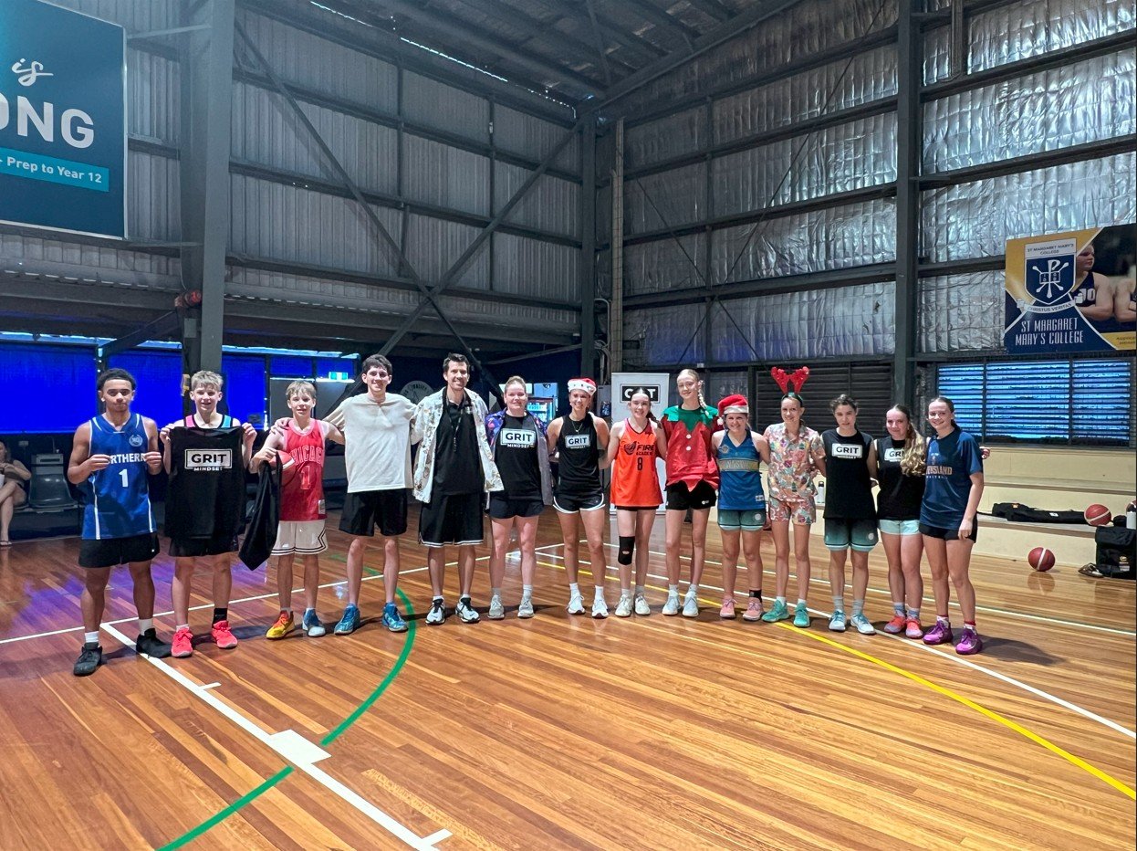 Group of 13 young women and men standing in a gymnasium, some wearing sports jerseys and others in holiday-themed accessories like Santa hats and reindeer antlers, posing for a photo on the basketball court.