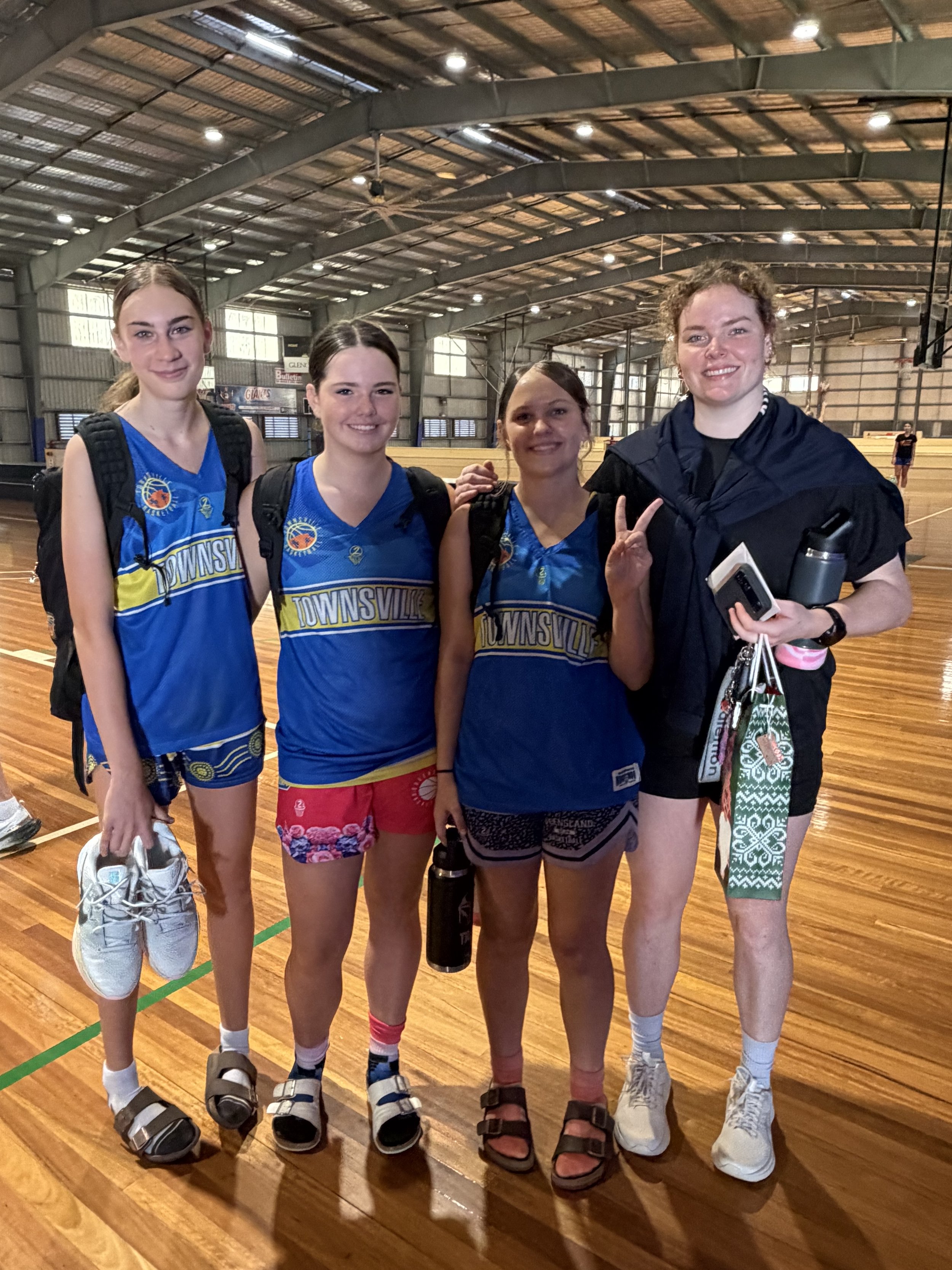 Four young women in sports uniforms and backpacks standing inside a gymnasium, smiling for a photo.