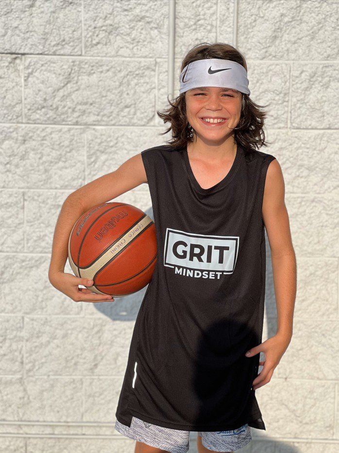 Young girl in a black basketball jersey with 'GRIT MINDSET' printed on it, holding a basketball, smiling, wearing a Nike headband, standing against a brick wall.