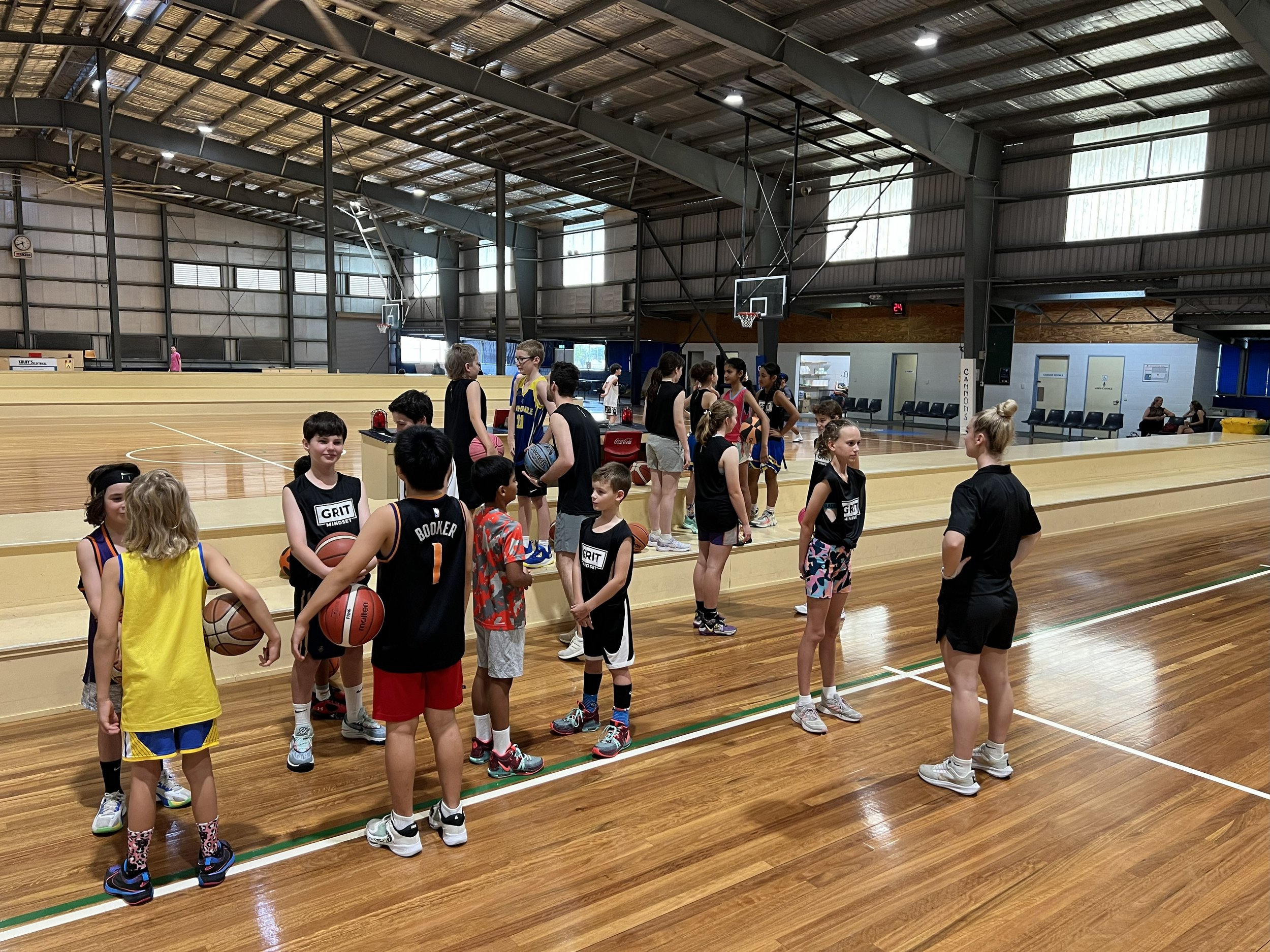 Children and teenagers gather in a gymnasium, some holding basketballs, likely for a basketball practice or class. There are two groups, one standing on the floor and the other on a raised platform. The gym has a polished wooden floor, high ceiling, 
