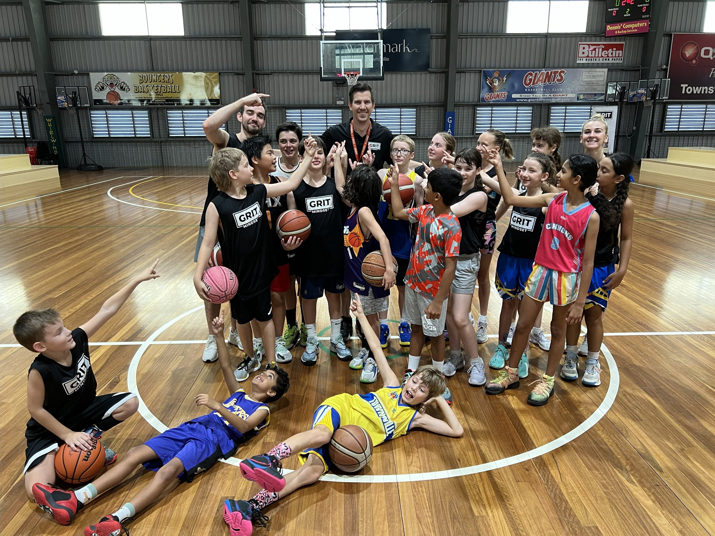 A group of children and two adult coaches on an indoor basketball court, celebrating after a youth basketball practice or game. The children are wearing basketball jerseys, holding basketballs, and some are seated or lying on the court, smiling and p