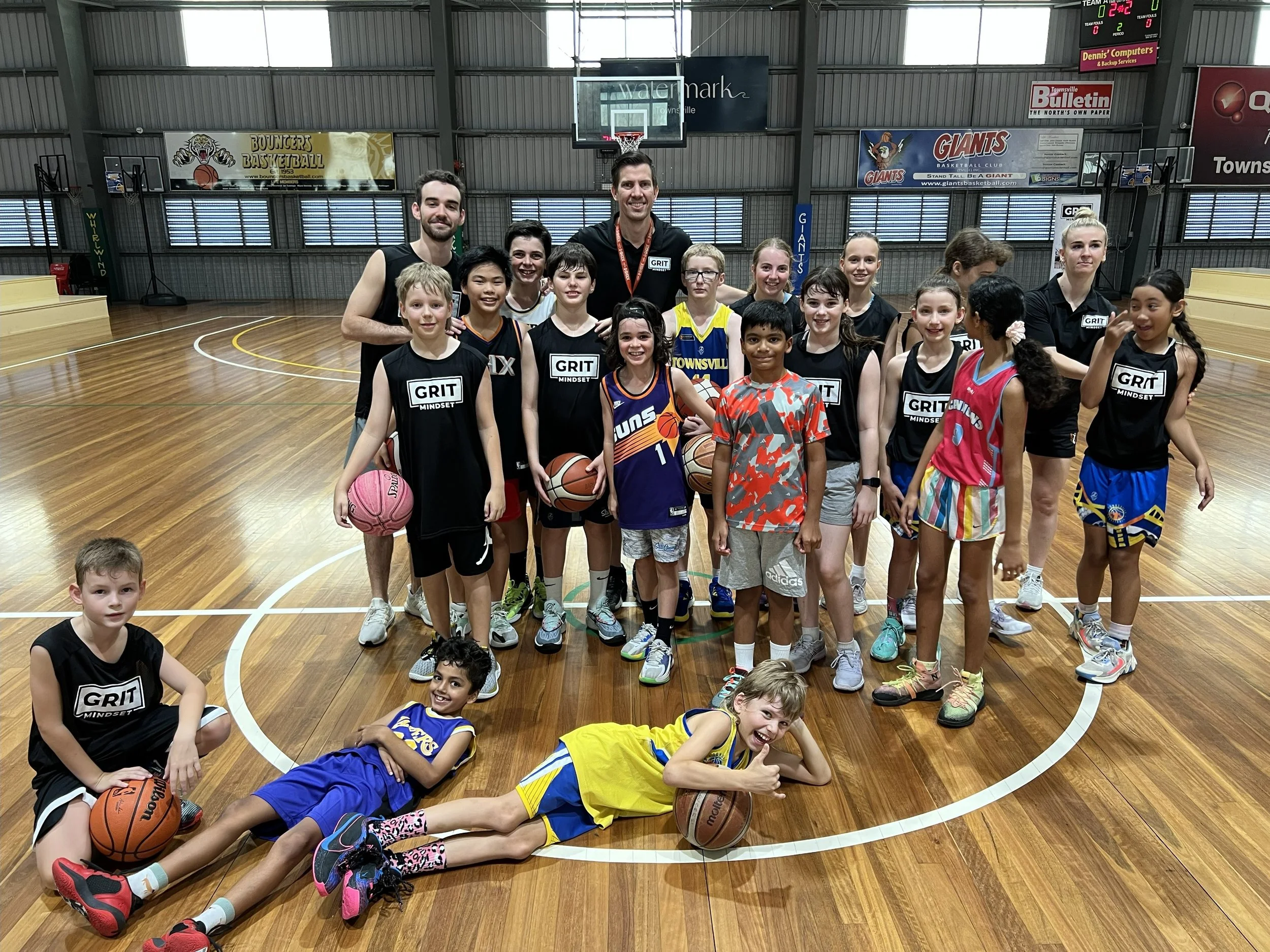 Group of young female and male basketball players, coaches, and children on an indoor basketball court, posing for a team photo. The players are wearing black jerseys with 