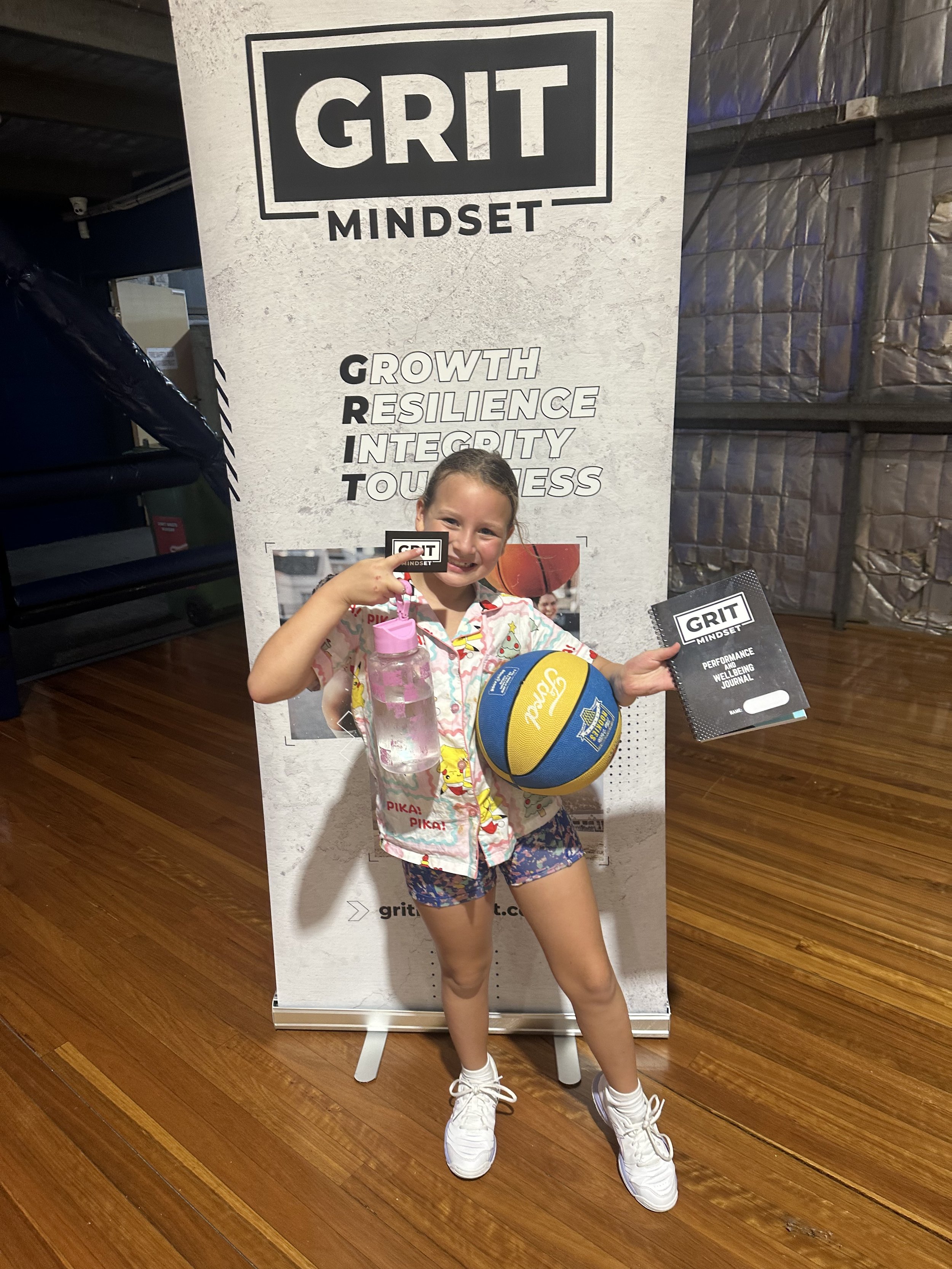 A young girl in colorful pajamas and shorts is smiling and holding a water bottle, a volleyball, a notebook, and a card in front of a Grit Mindset promotional banner. The banner lists qualities like growth, resilience, integrity, and toughness.
