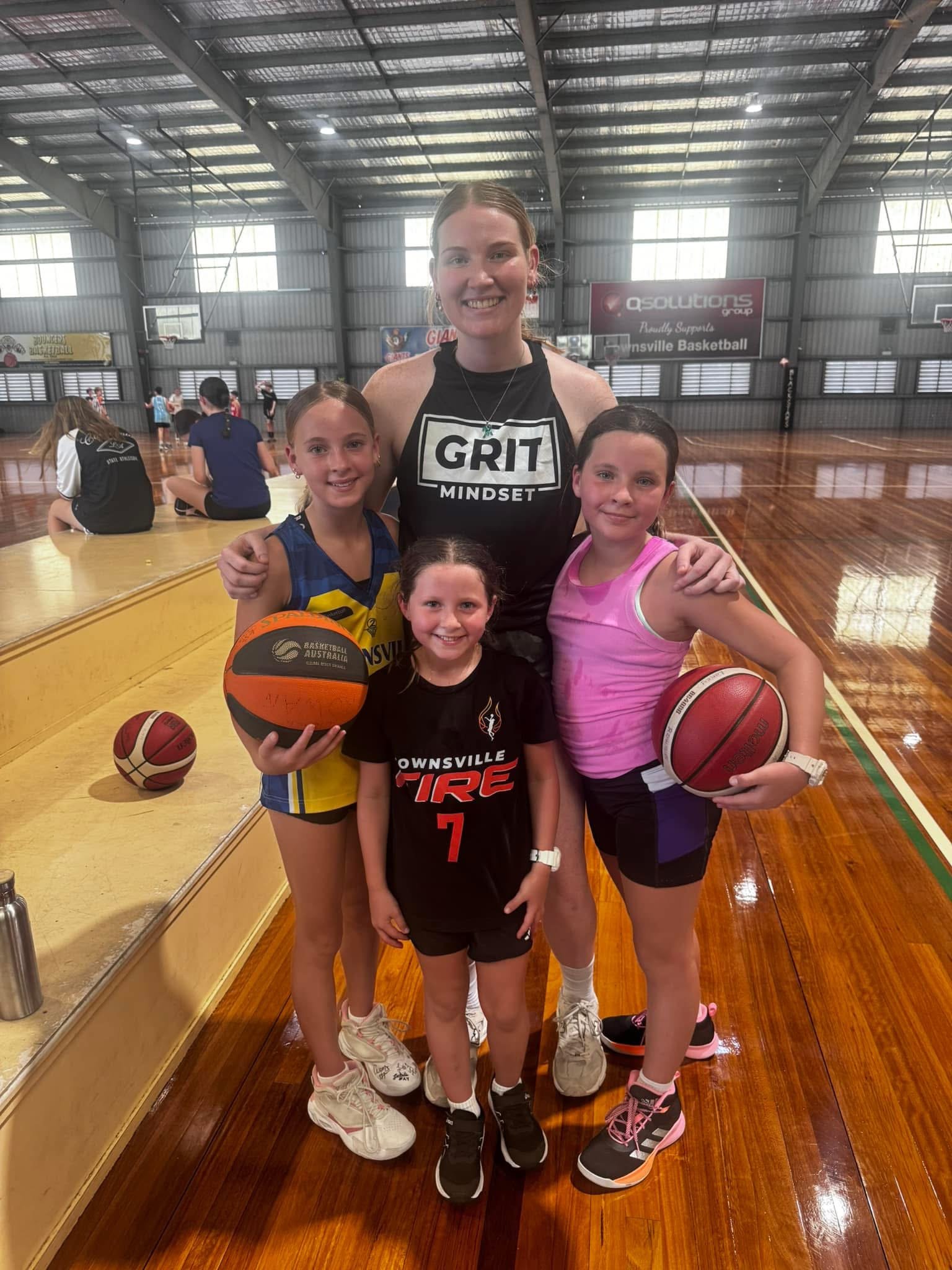 A group of young girls and a woman basketball coach inside a gymnasium, with wooden flooring, holding basketballs, smiling for a photo.
