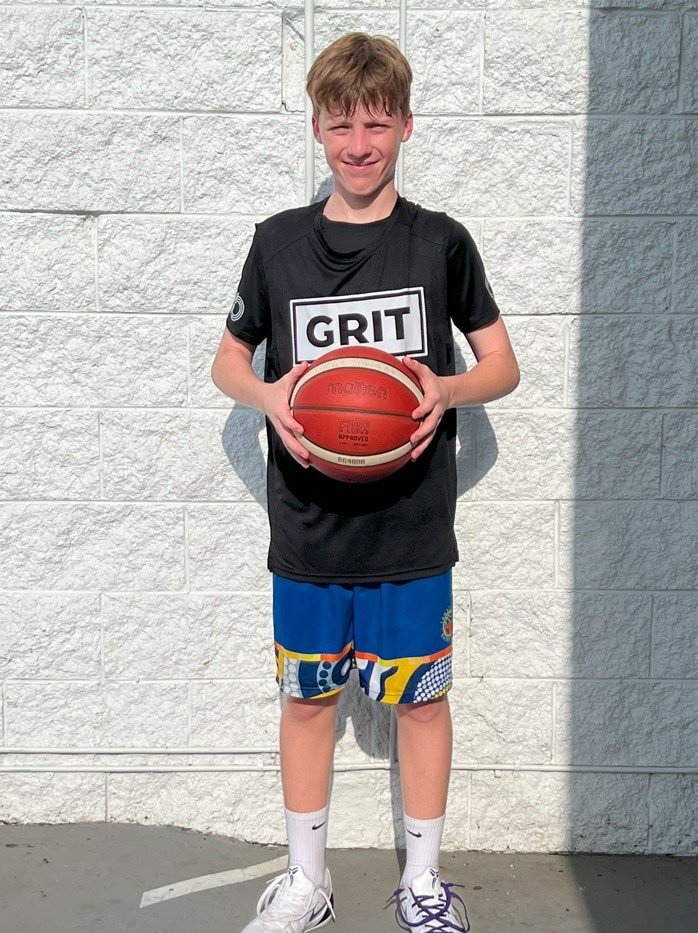 A young boy standing outdoors against a white brick wall, holding a red basketball, wearing a black T-shirt with the word "GRIT" on it, colorful shorts, white socks, and white sneakers.