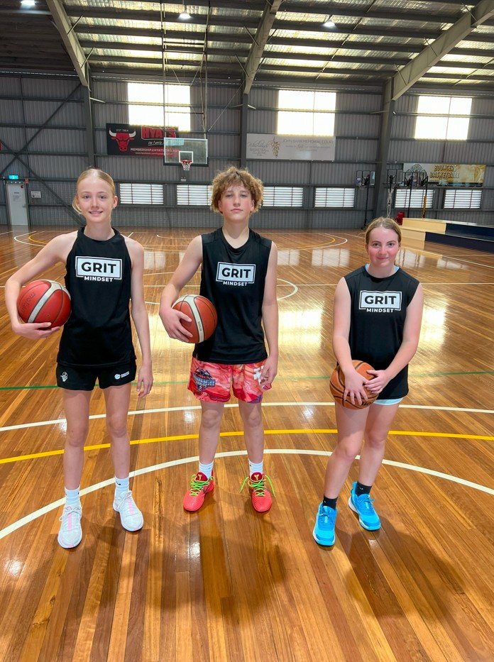 Three young girls in basketball uniforms holding basketballs on an indoor basketball court.
