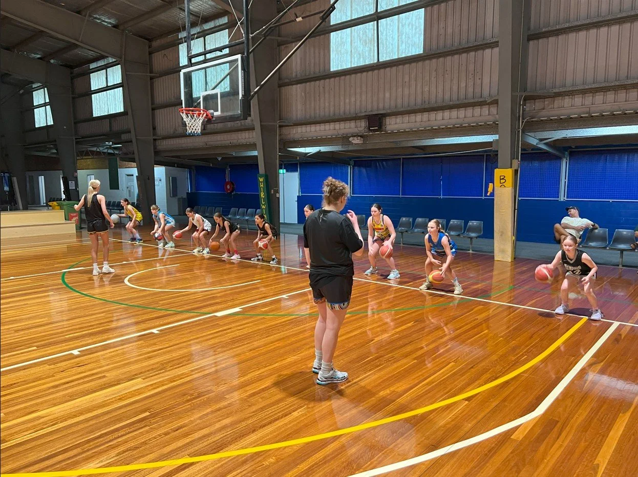 A basketball coach instructing young players during a practice on a hardwood indoor court, with some seated spectators in the background.