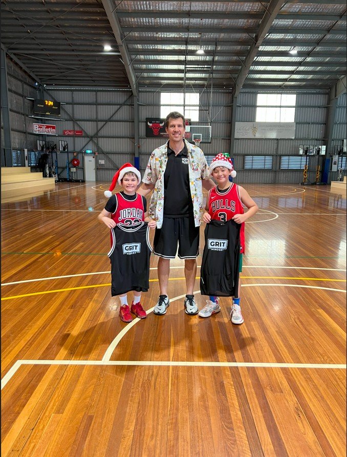 Two children dressed in Chicago Bulls basketball jerseys with Santa hats, standing on a basketball court with a man in casual clothing in the middle, inside a sports gymnasium.