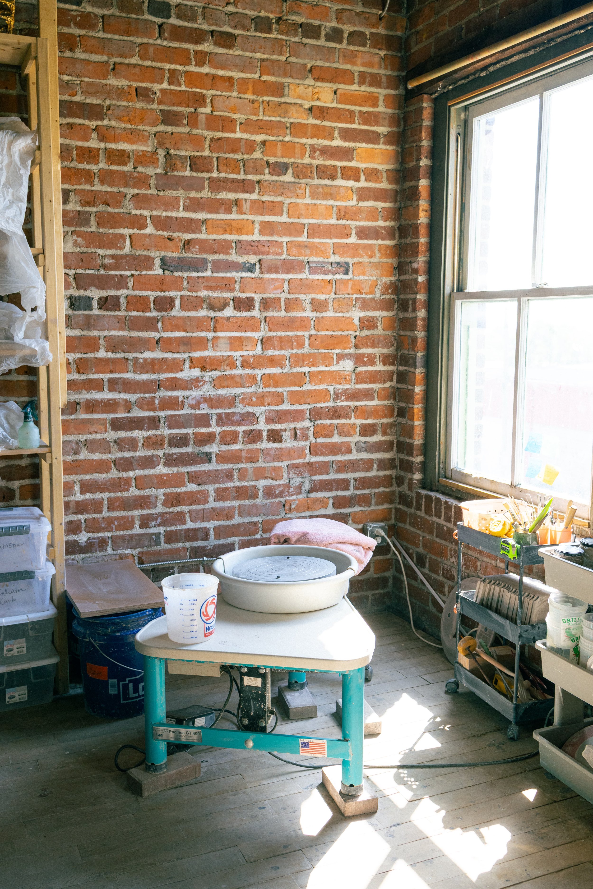 A sunny pottery studio with a brick wall, a window on the right, and various tools and supplies on shelves and tables, including a pottery wheel, and clay crafting supplies.