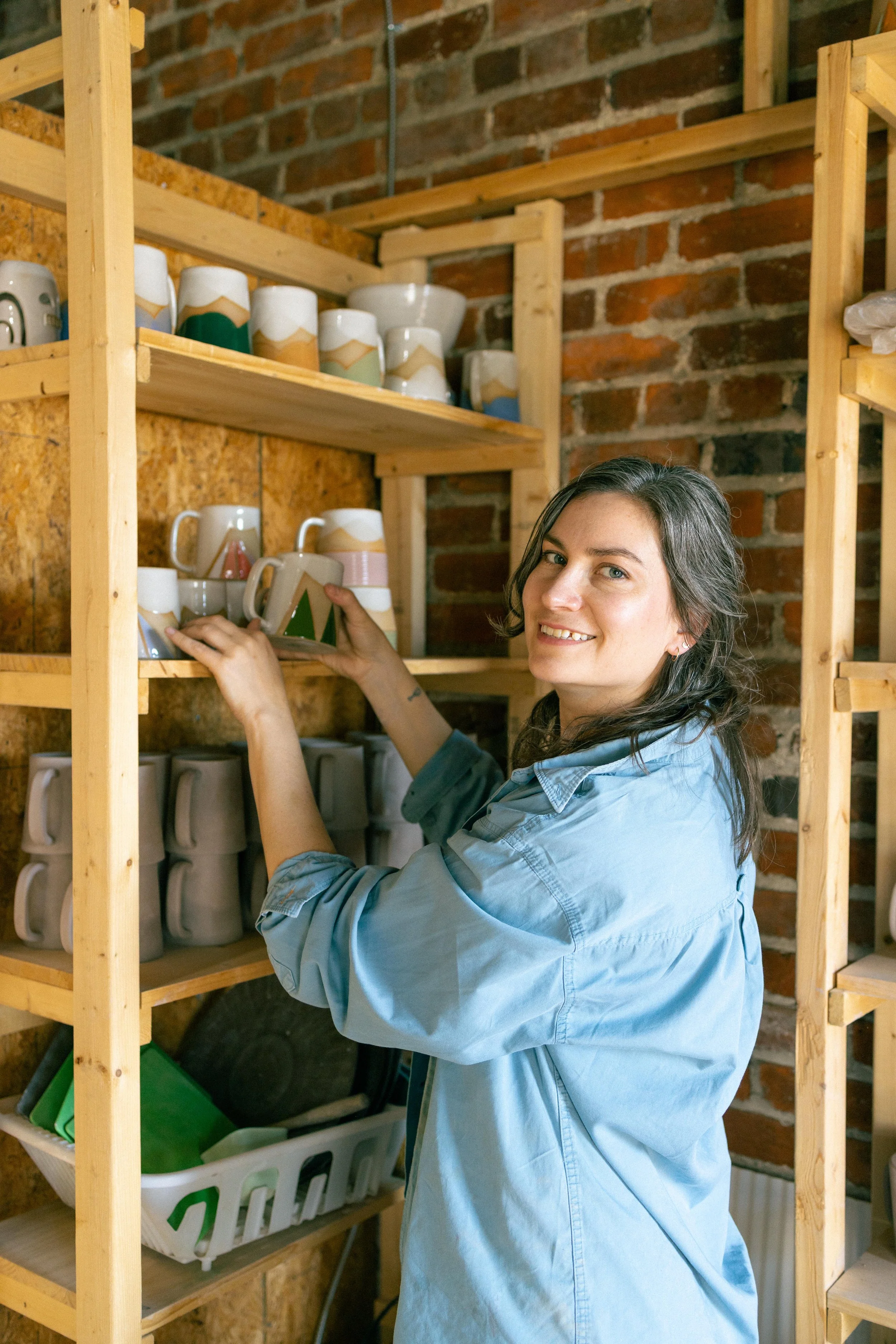 A woman with dark hair and a blue shirt organizing ceramic mugs on a wooden shelf against a brick wall.