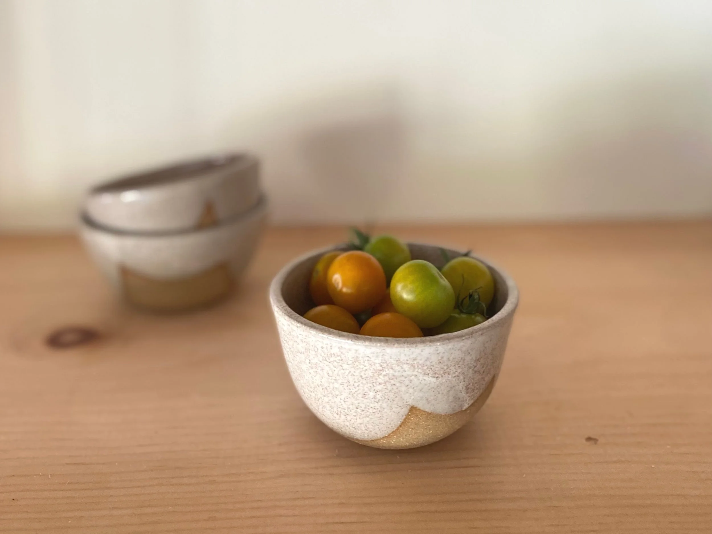 A small ceramic bowl filled with yellow and green cherry tomatoes on a wooden surface, with two white bowls in the background.
