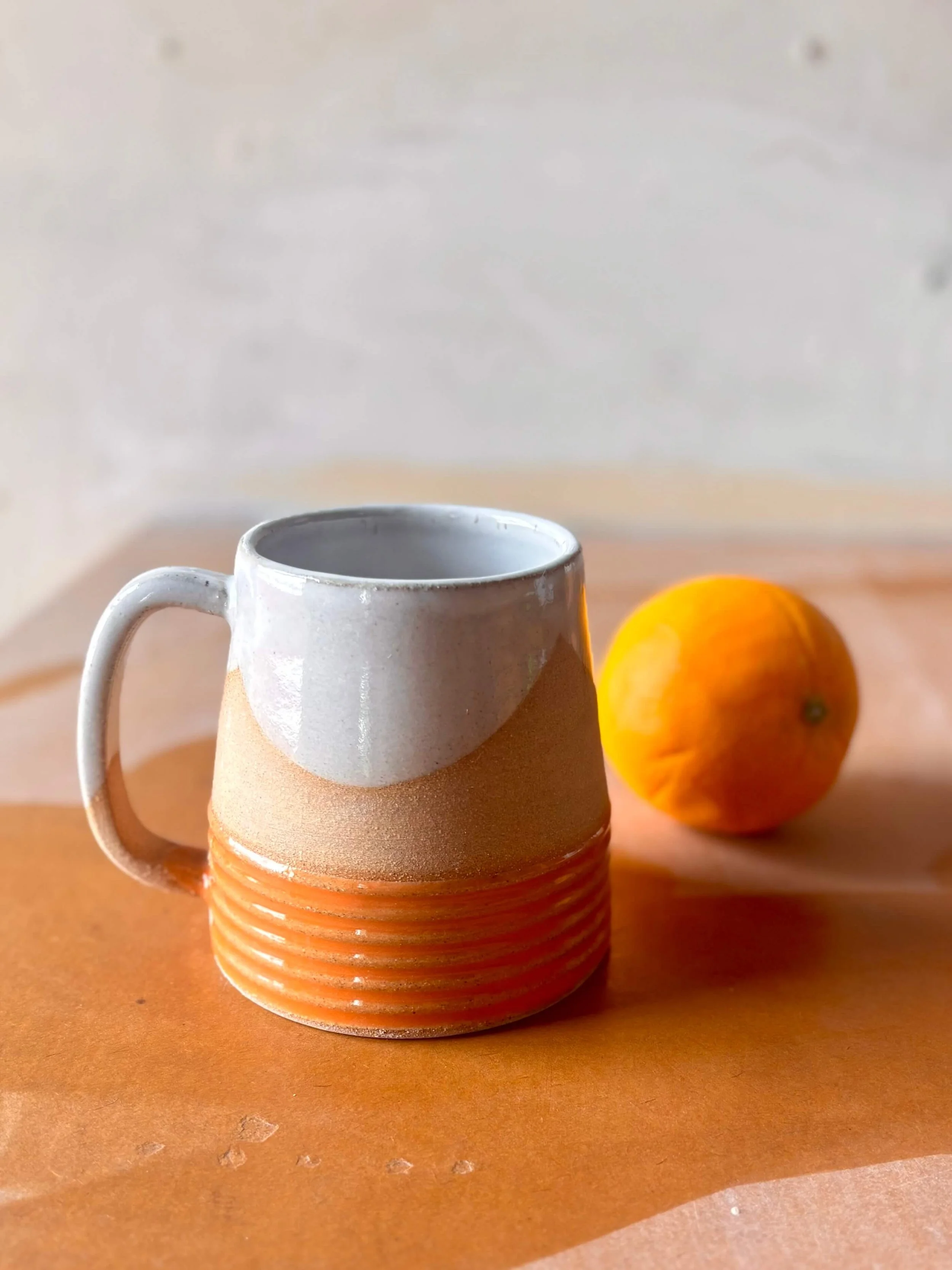 A ceramic mug with a white upper half and an orange lower half and a fluted texture on the lower third, alongside an orange, on a wooden surface.