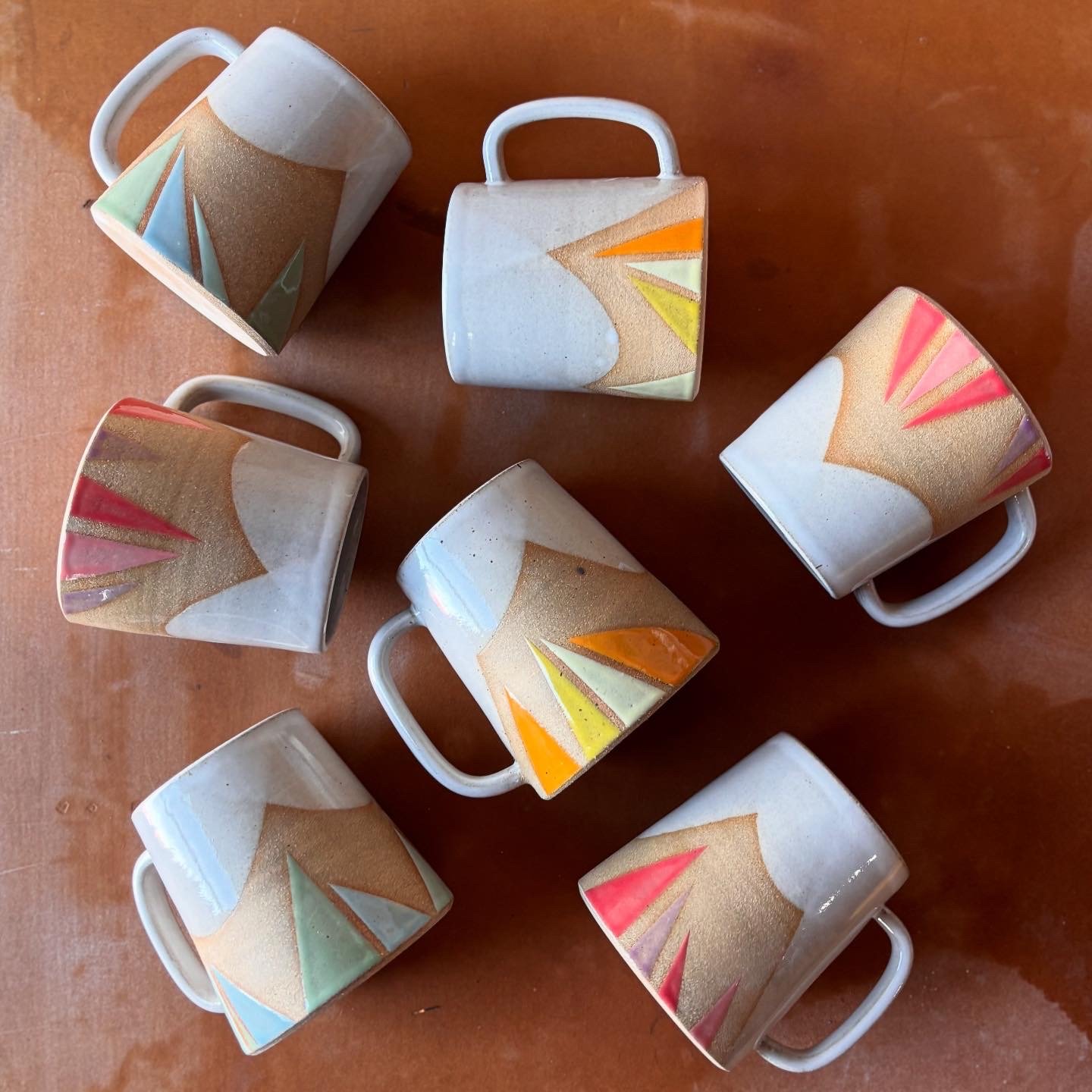 Six ceramic mugs with geometric designs in pastel colors, placed on a wooden surface.