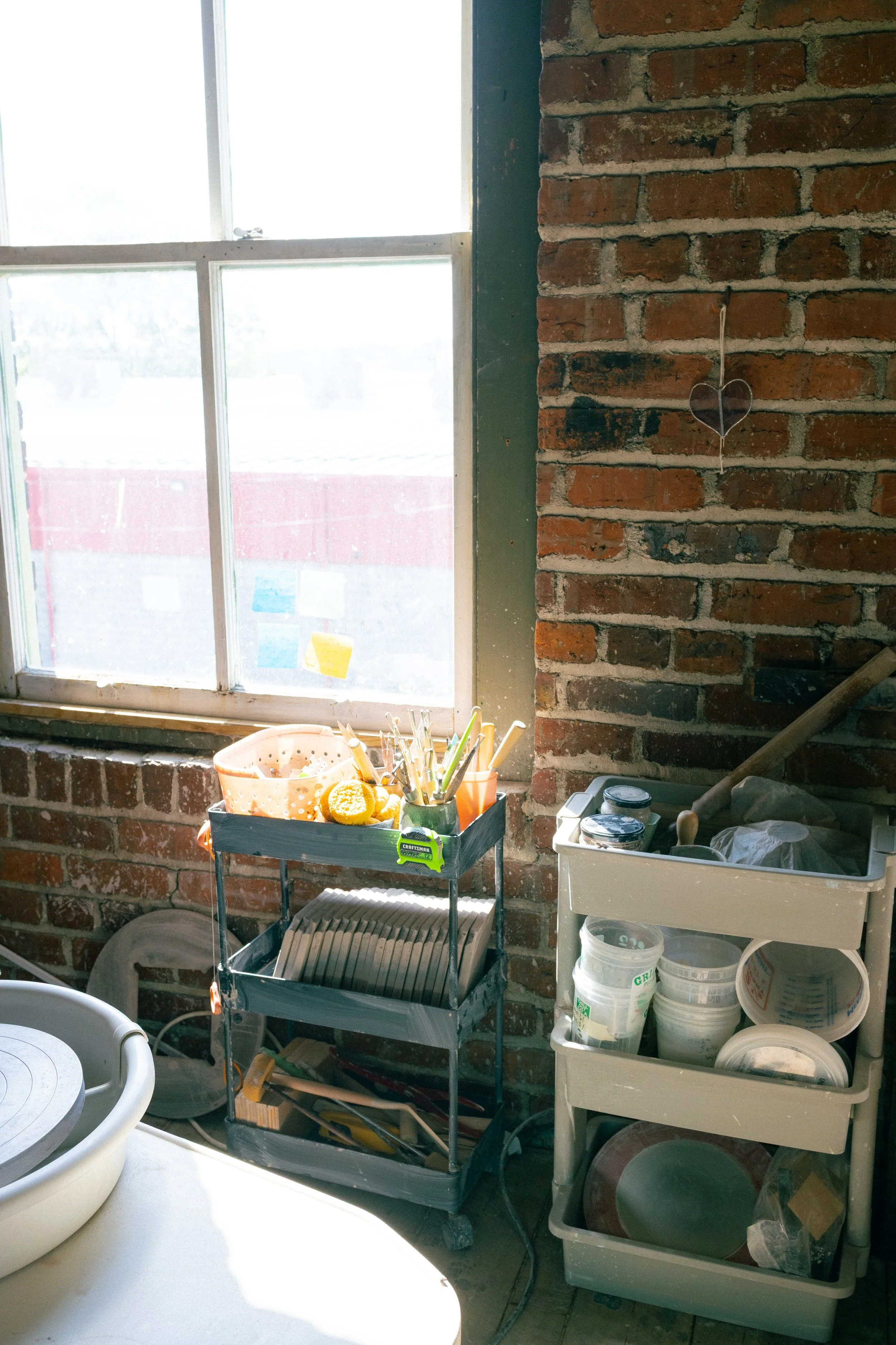 A windowsill with various pottery tools, containers, and supplies in a room with a brick wall. There is a decorative heart hanging on the brick wall and sunlight coming through the window.