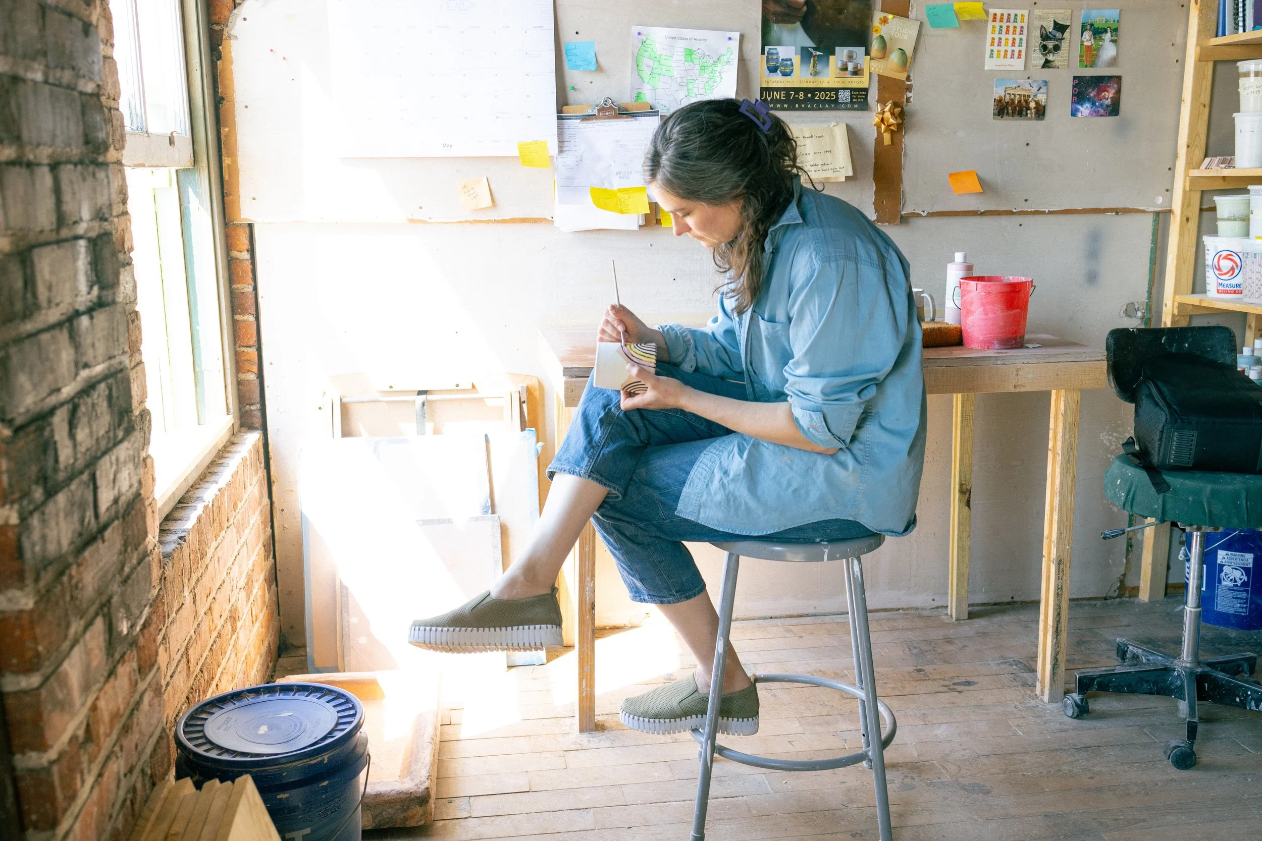 A woman sitting on a high stool with one foot resting on a rung, glazing pottery in a room with a brick wall on the left and a bulletin board with various paper notes and pictures on the wall. Sunlight is coming in from a window to the left.