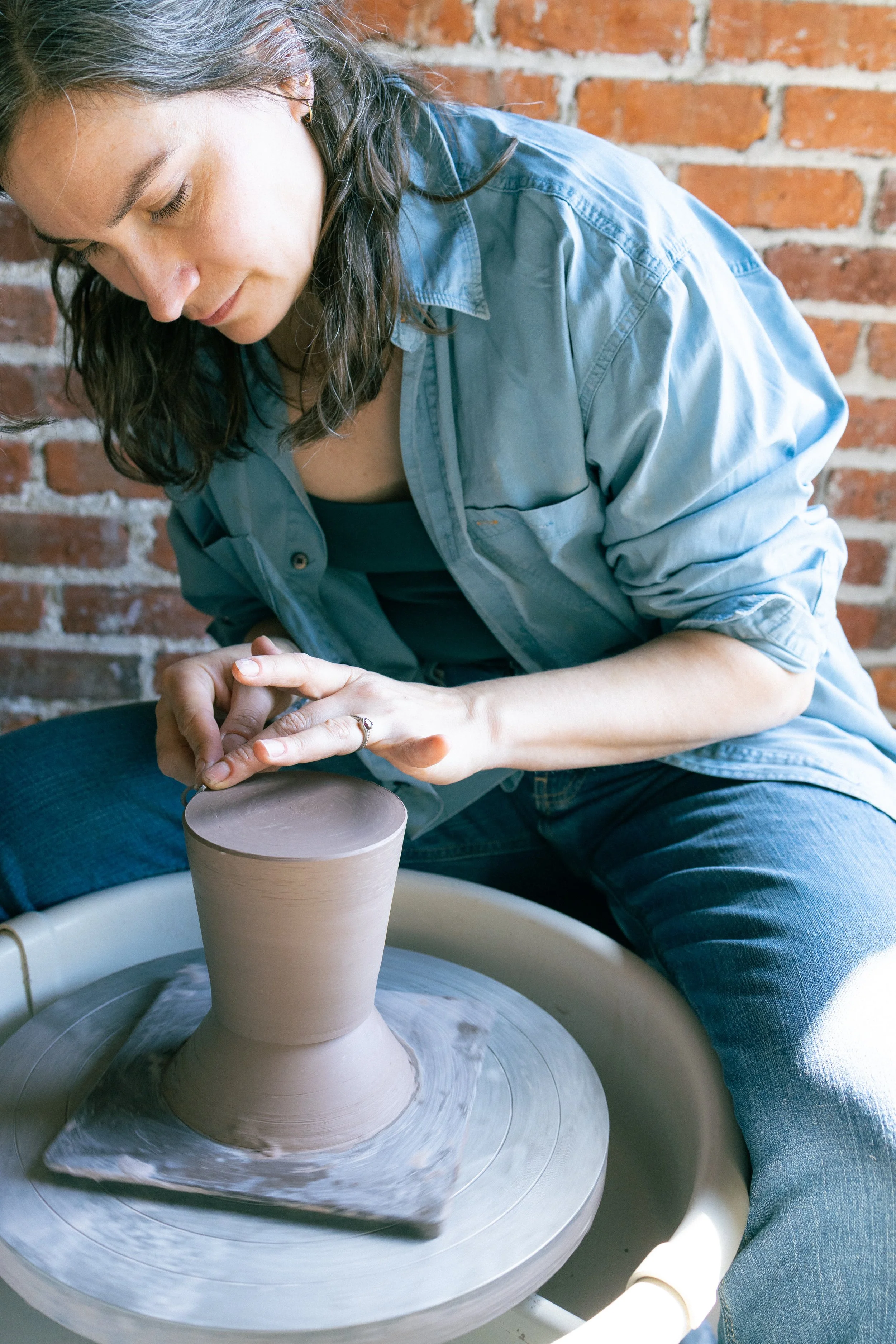 A woman sculpting pottery on a wheel in a studio with a brick wall background.