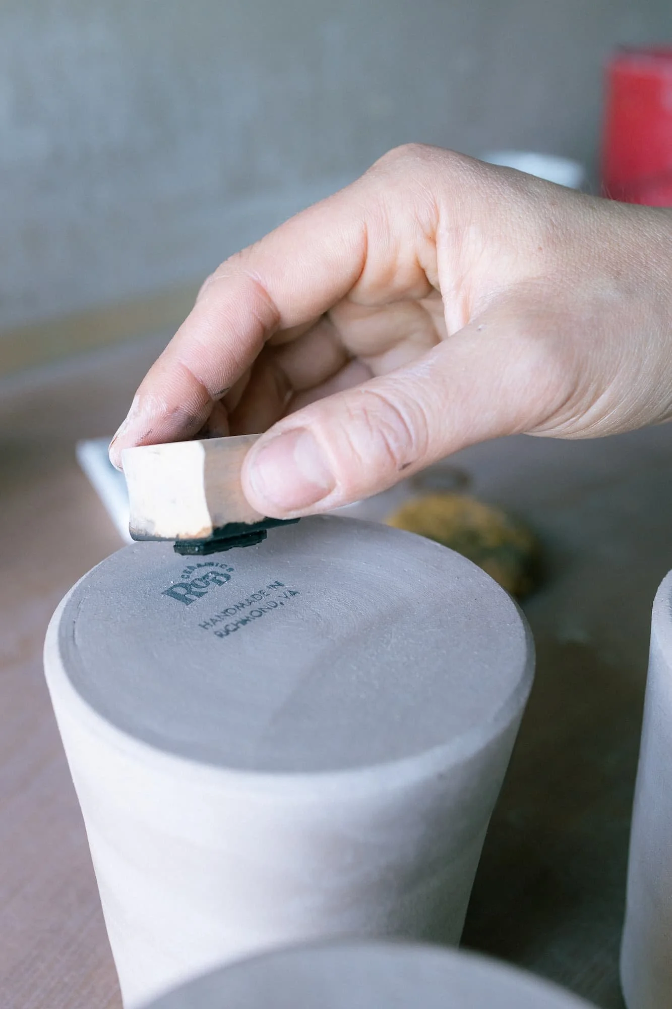 Close-up of a hand holding a small ROB Ceramics maker's mark stamp in pottery studio setting.