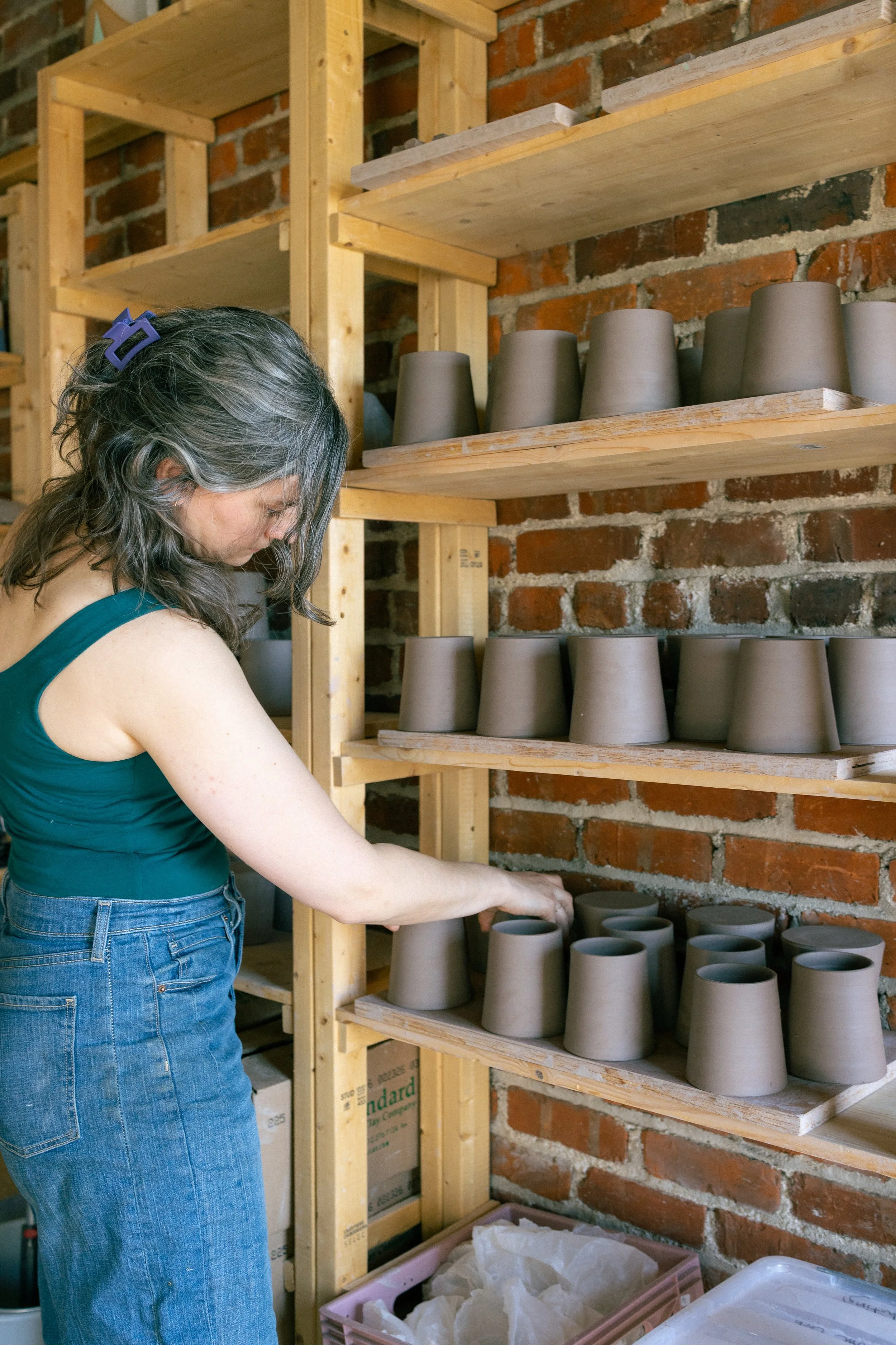 A woman with gray hair and a blue hair clip working with unpainted ceramic pots on wooden shelves in a pottery studio with a brick wall background.