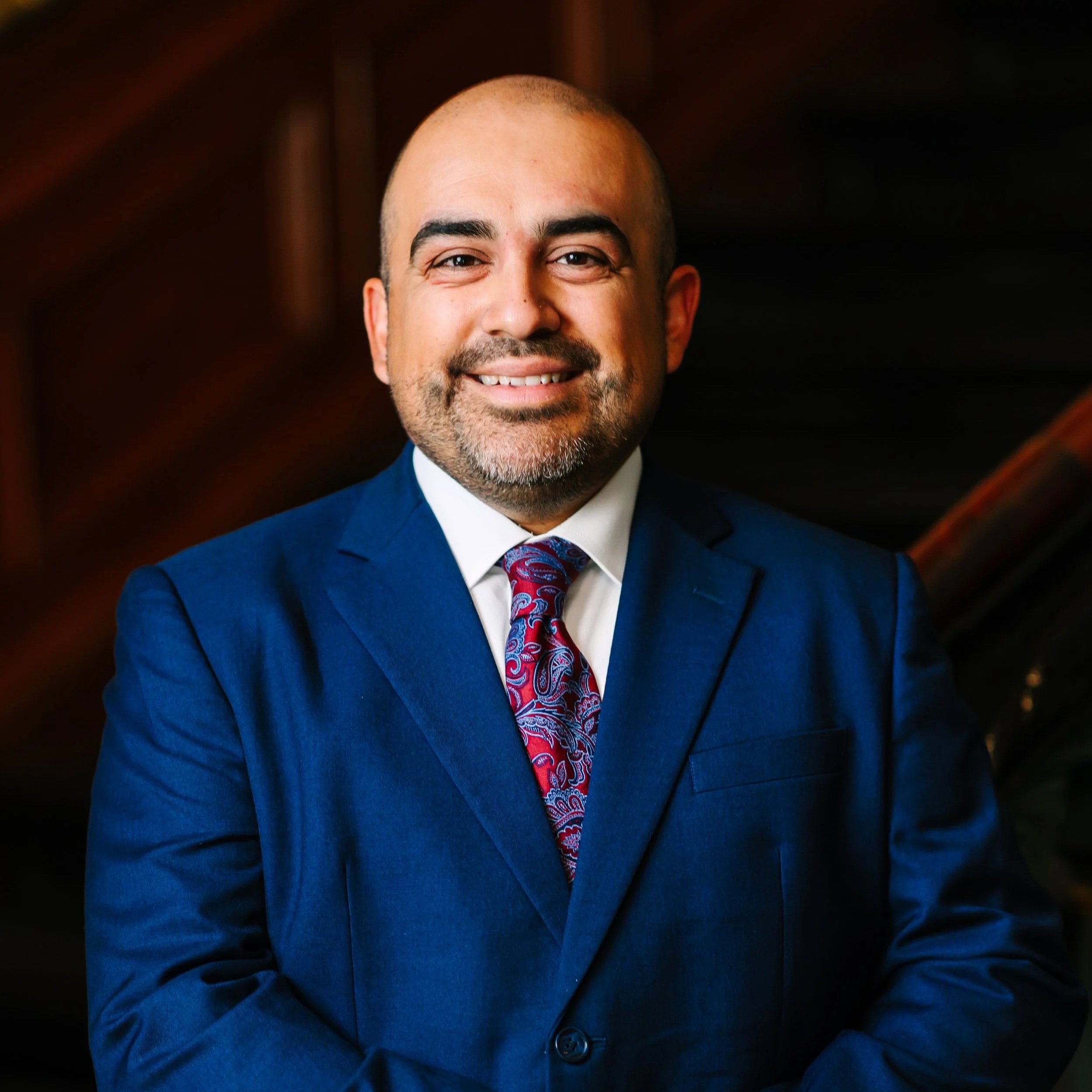 Headshot of a man with a shaved head, beard, and mustache, wearing a blue suit jacket and a patterned red and blue tie, smiling against a dark background.