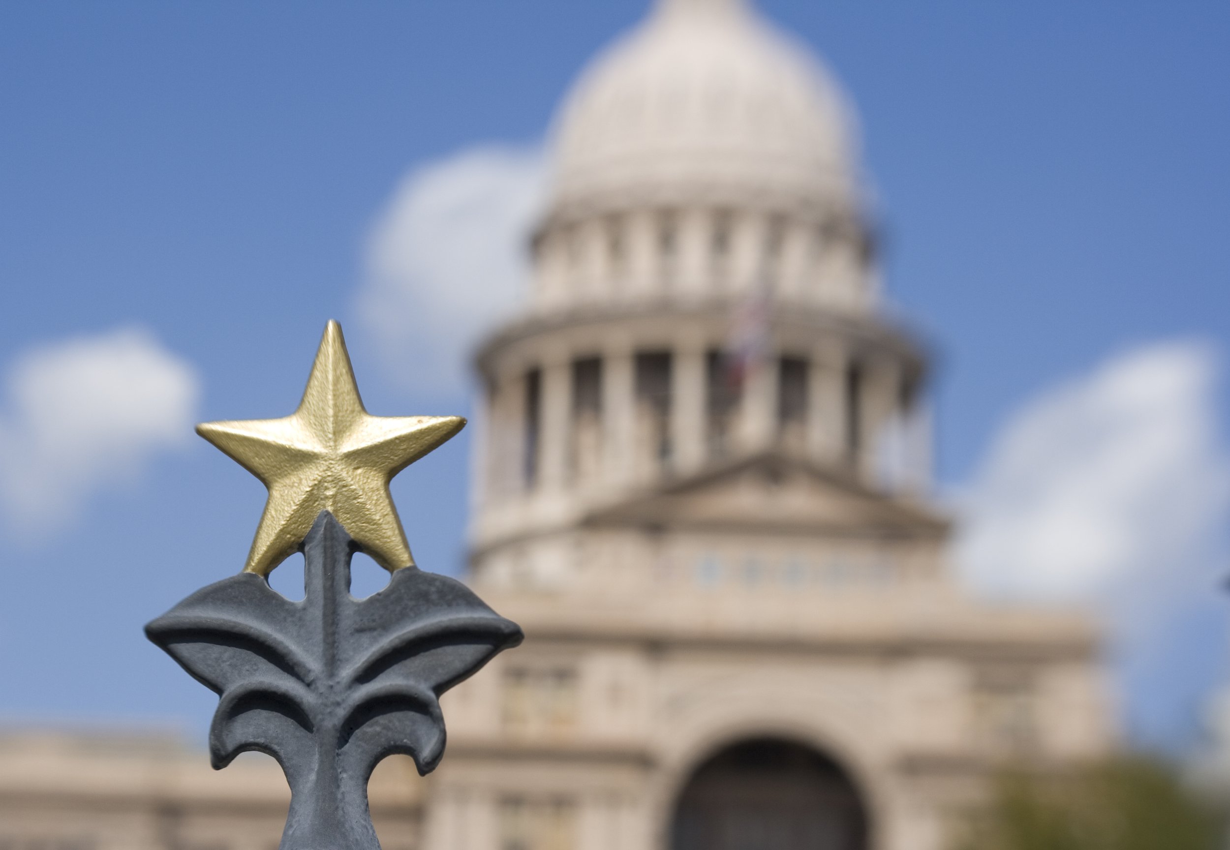 Close-up of a decorative fence post with gold and gray stars, with the United States Capitol building blurred in the background against a blue sky with clouds.