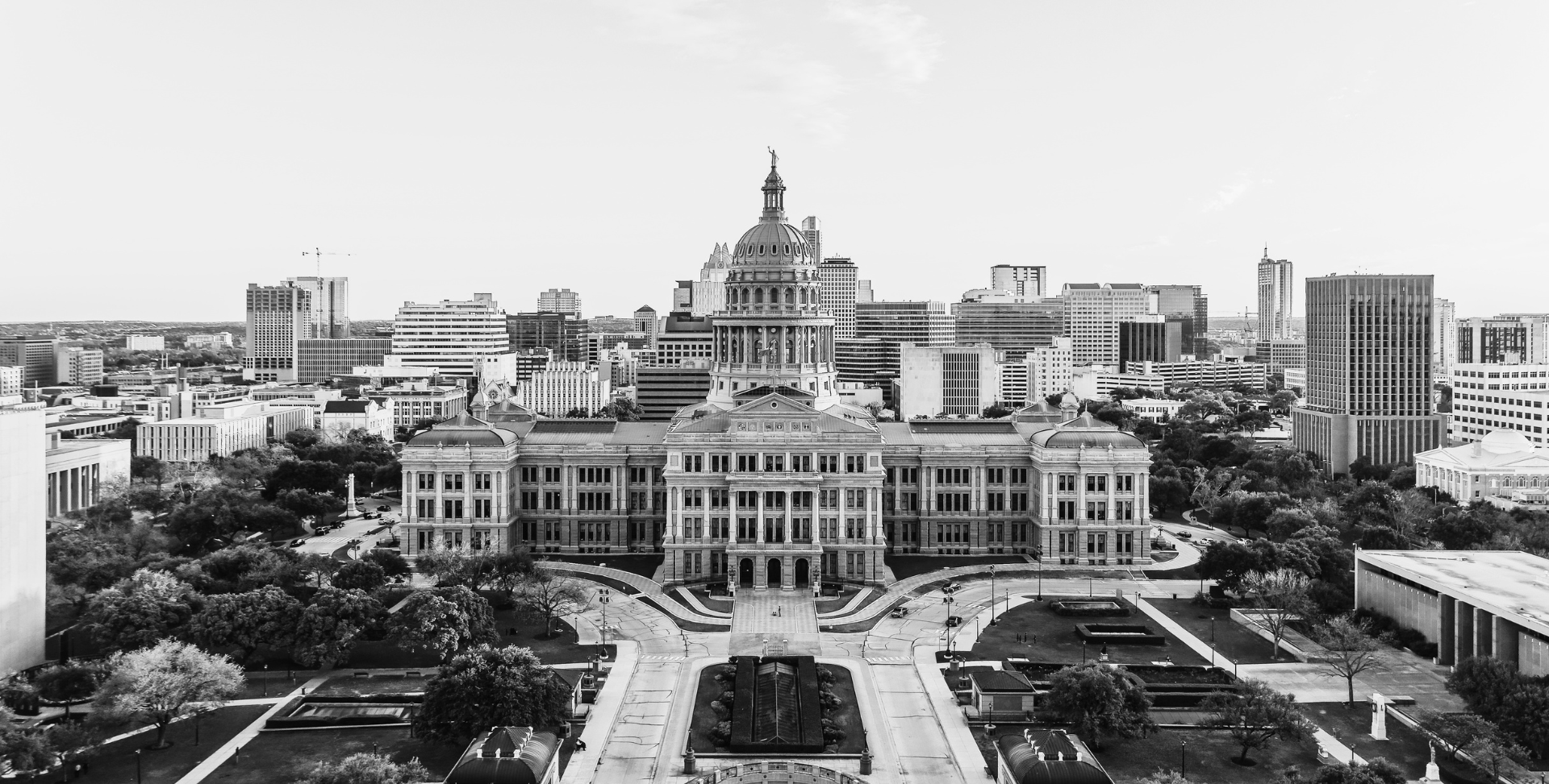 Black and white aerial view of a historic government building with a dome and a statue on top, surrounded by modern skyscrapers and trees in an urban cityscape.