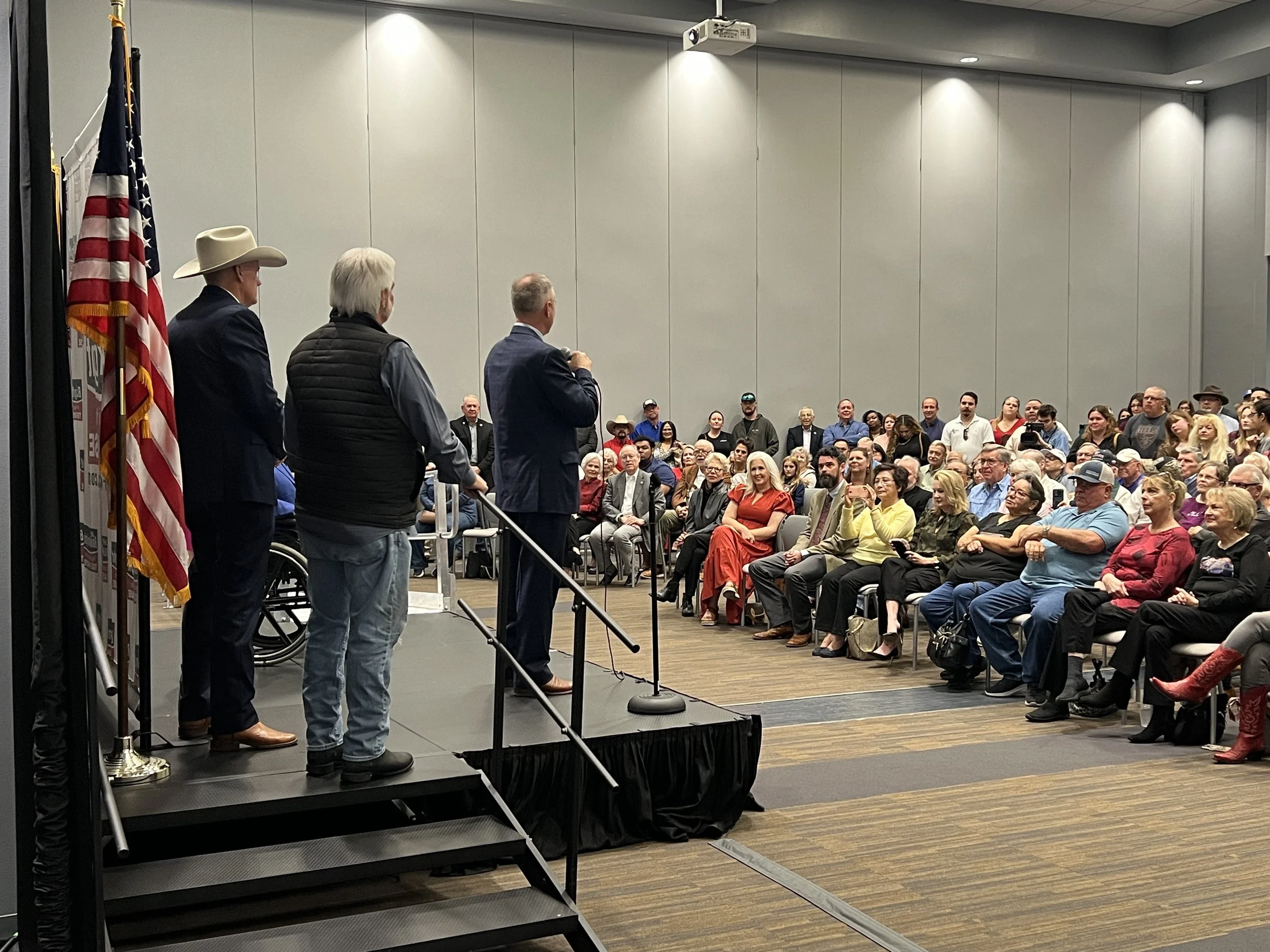 A speaker is addressing a seated audience at a conference, with three men standing on the stage near the speaker, and an American flag positioned near the stage.