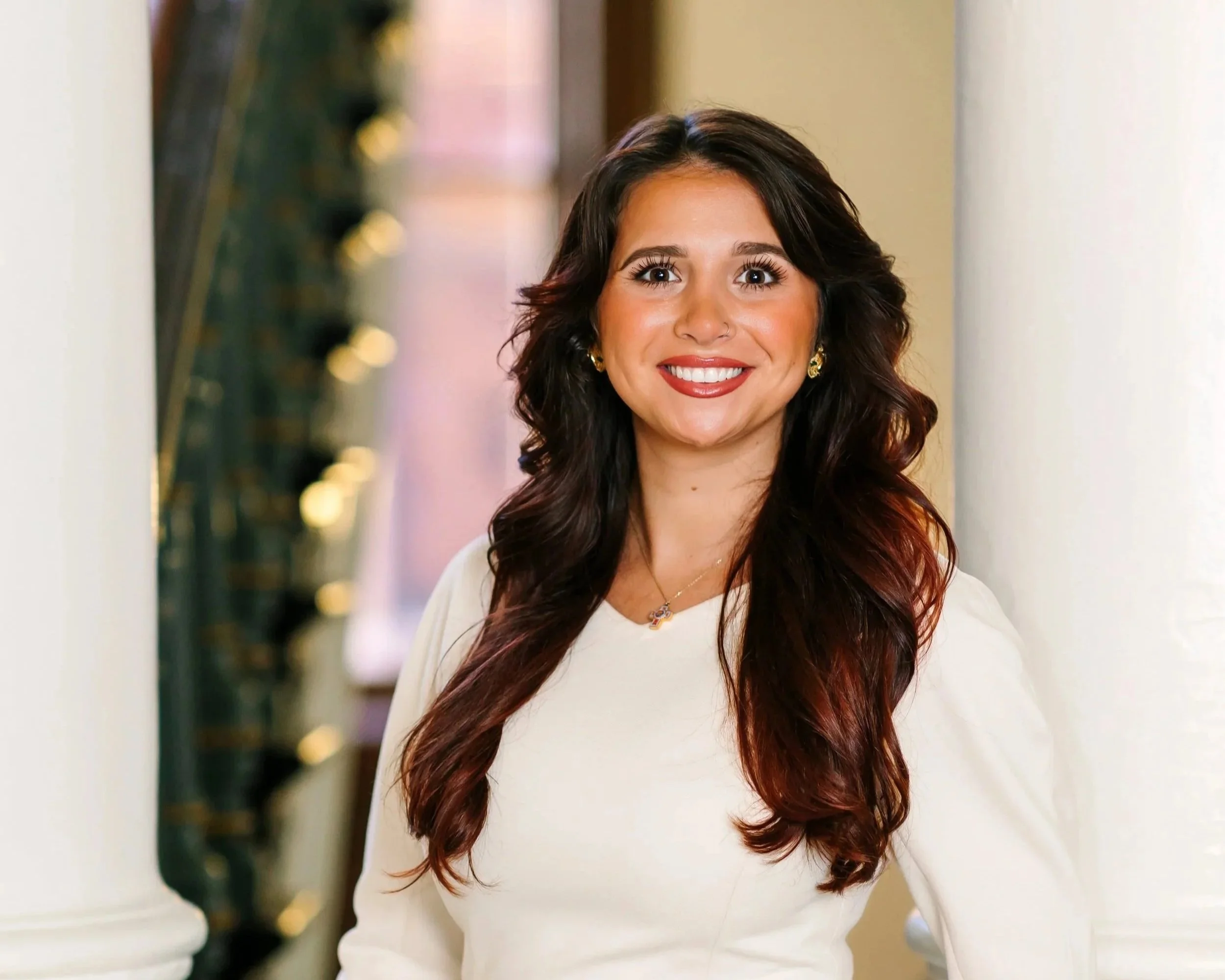 Portrait of a woman with long, wavy dark brown and red hair, smiling, wearing gold earrings and a white top.