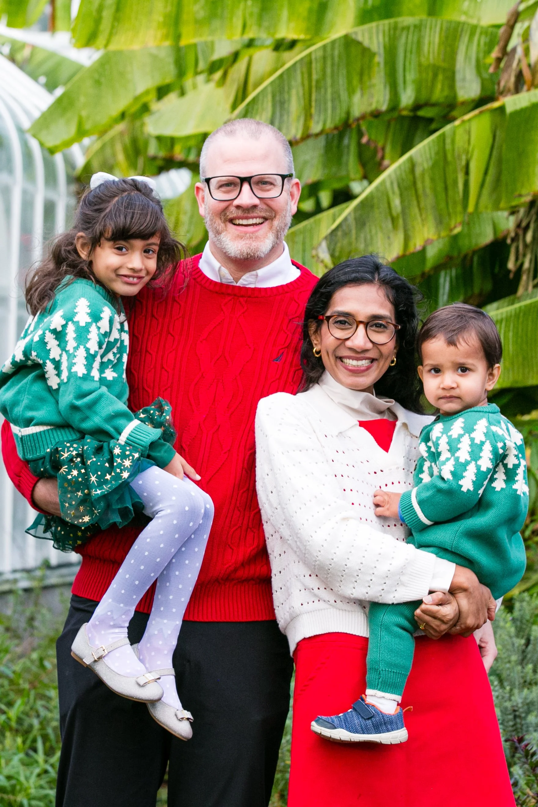 A happy family of four posing outdoors surrounded by large green banana leaves, with two children, a man, and a woman smiling at the camera.
