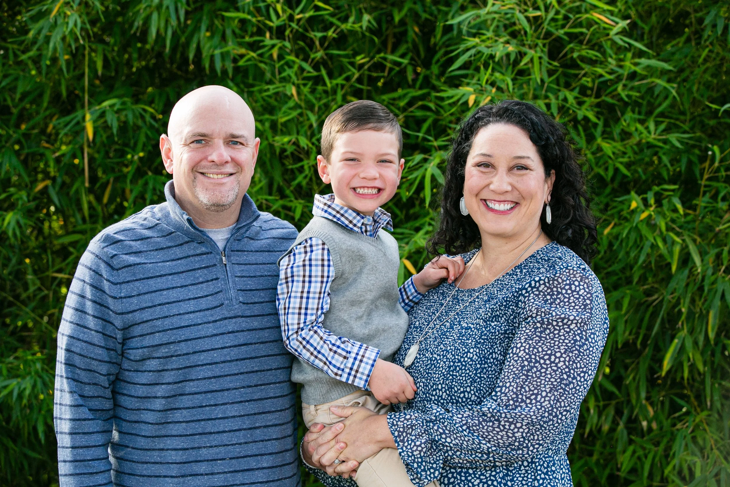 Happy family of three outdoors in front of green foliage, consisting of a bald man with a beard in a blue striped sweater, a smiling young boy in a gray vest and checkered shirt, and a woman with curly dark hair in a blue patterned dress holding the 