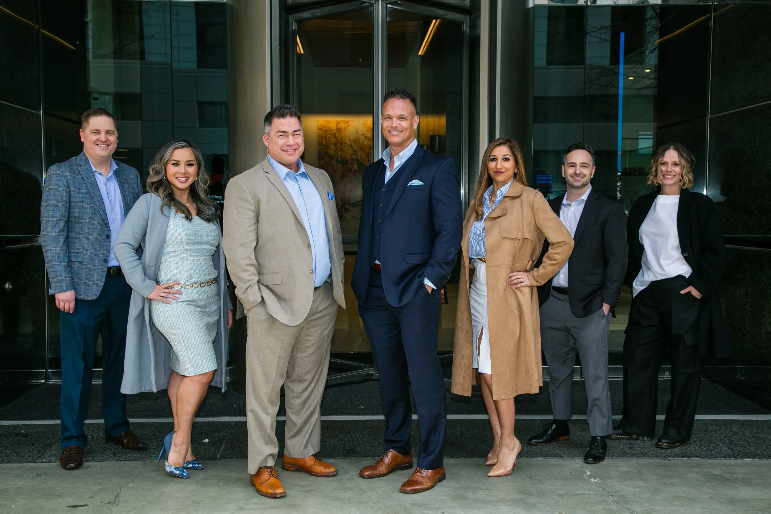 Group of seven diverse professionals standing outside a building, posing for a photo. All are dressed in business attire and smiling.
