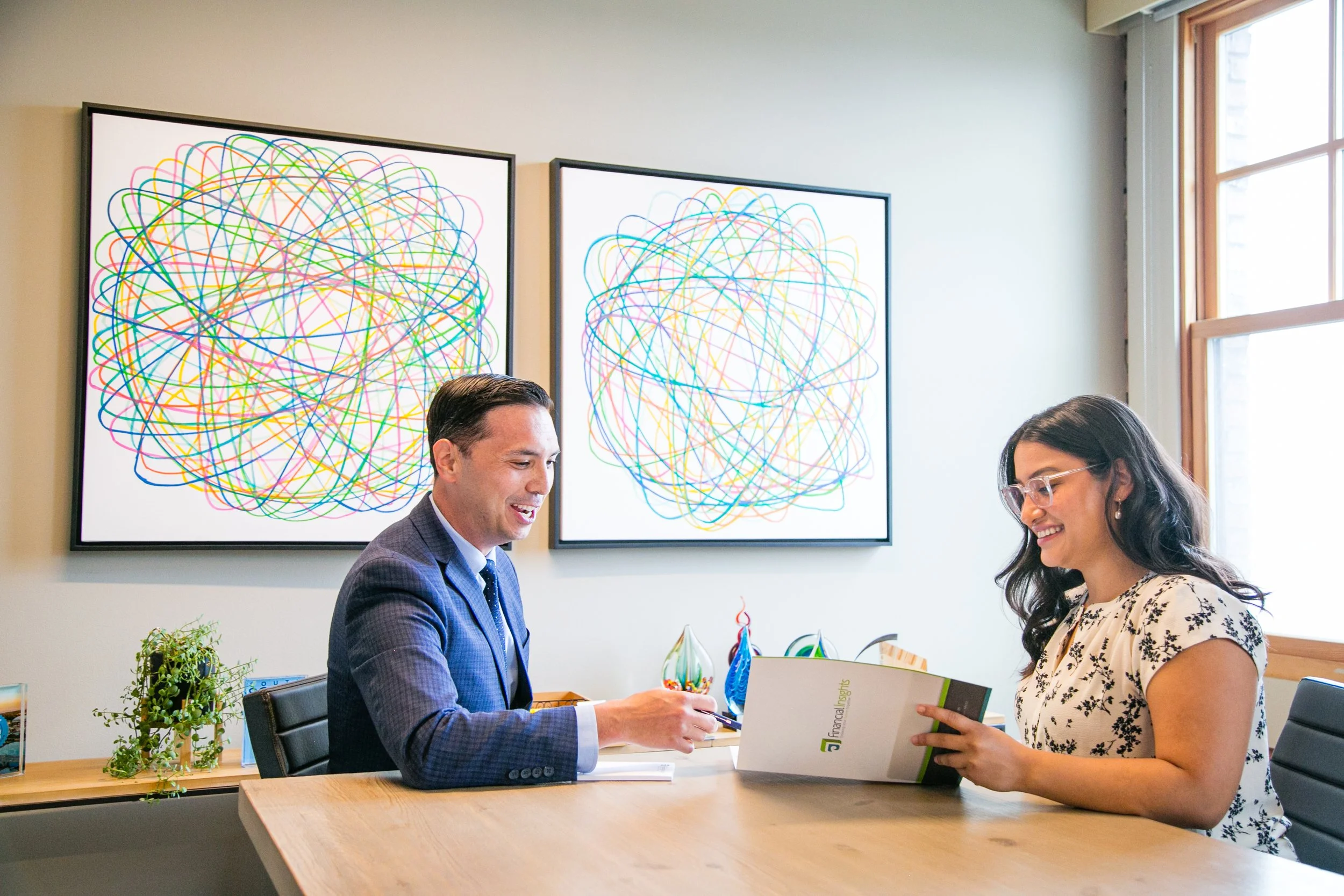 A man and woman sitting at a office table, smiling and looking at a document, with abstract colorful art on the wall behind them and a window to the right.