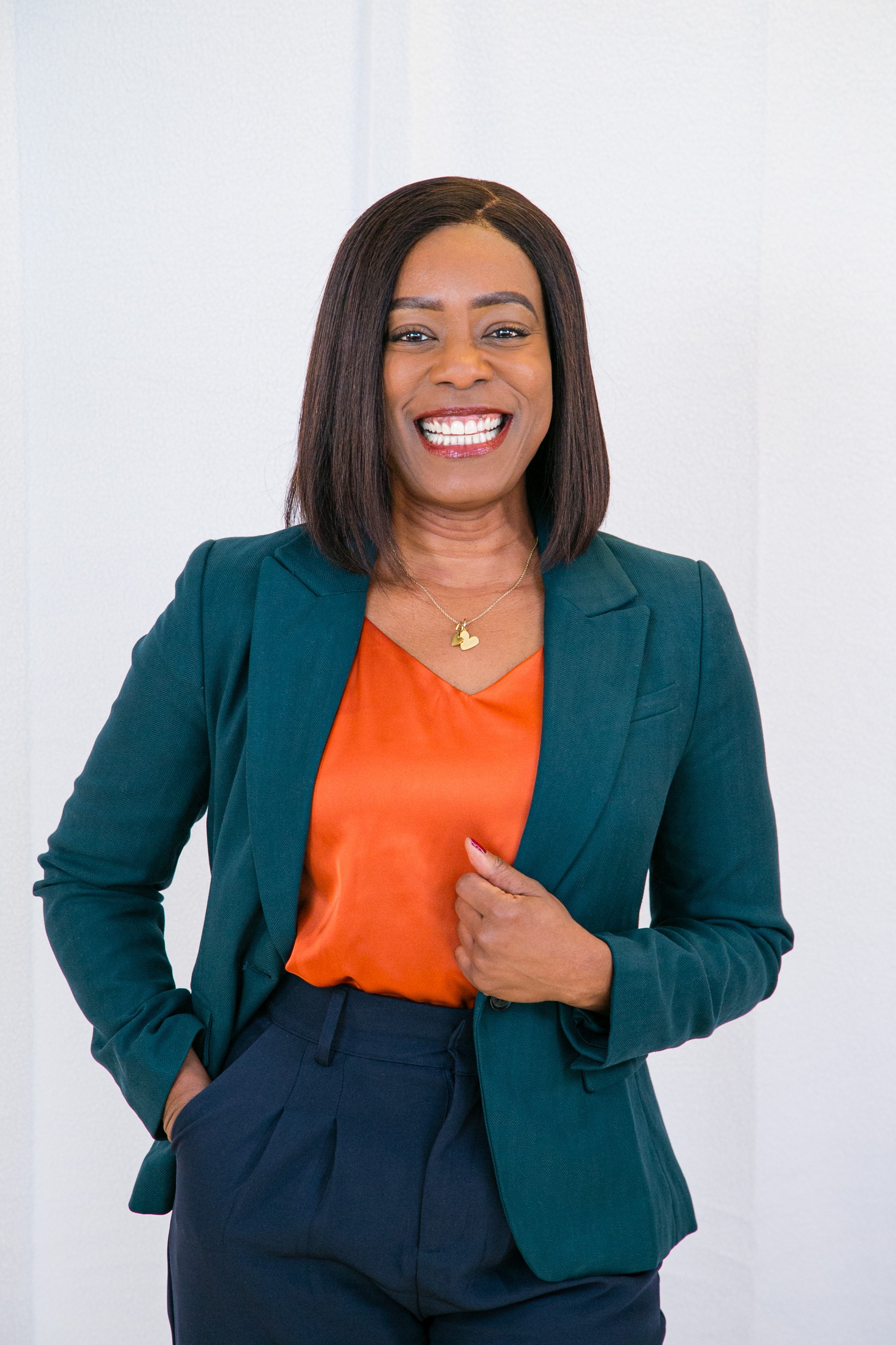 A smiling African American woman with shoulder-length dark hair, wearing a dark green blazer over an orange blouse, and dark green pants, standing against a plain white wall.