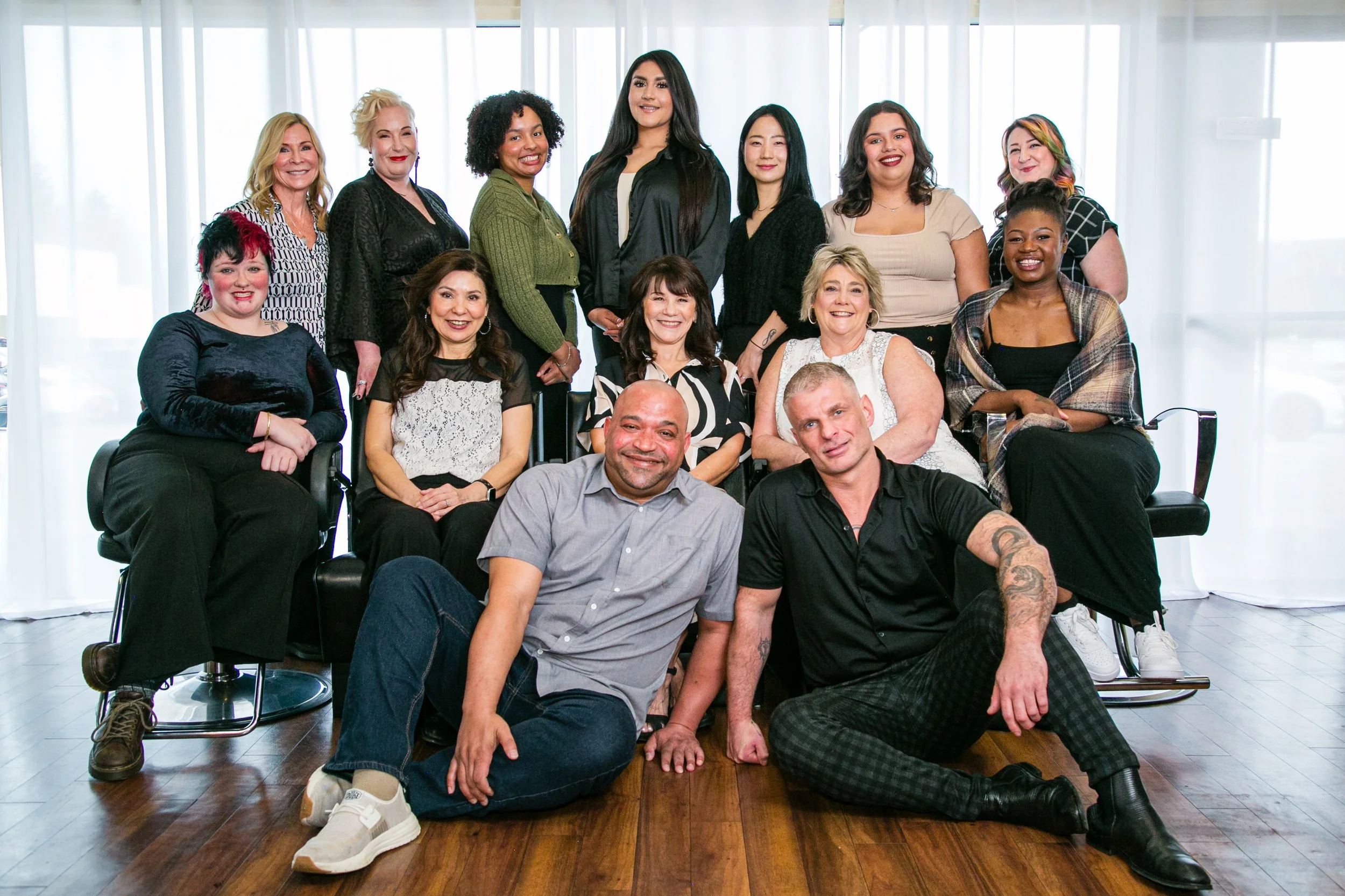 A diverse group of 14 adults, including men and women of various ethnicities, posing together indoors with large windows and white curtains in the background. Some are sitting, some are standing, and they are smiling at the camera.