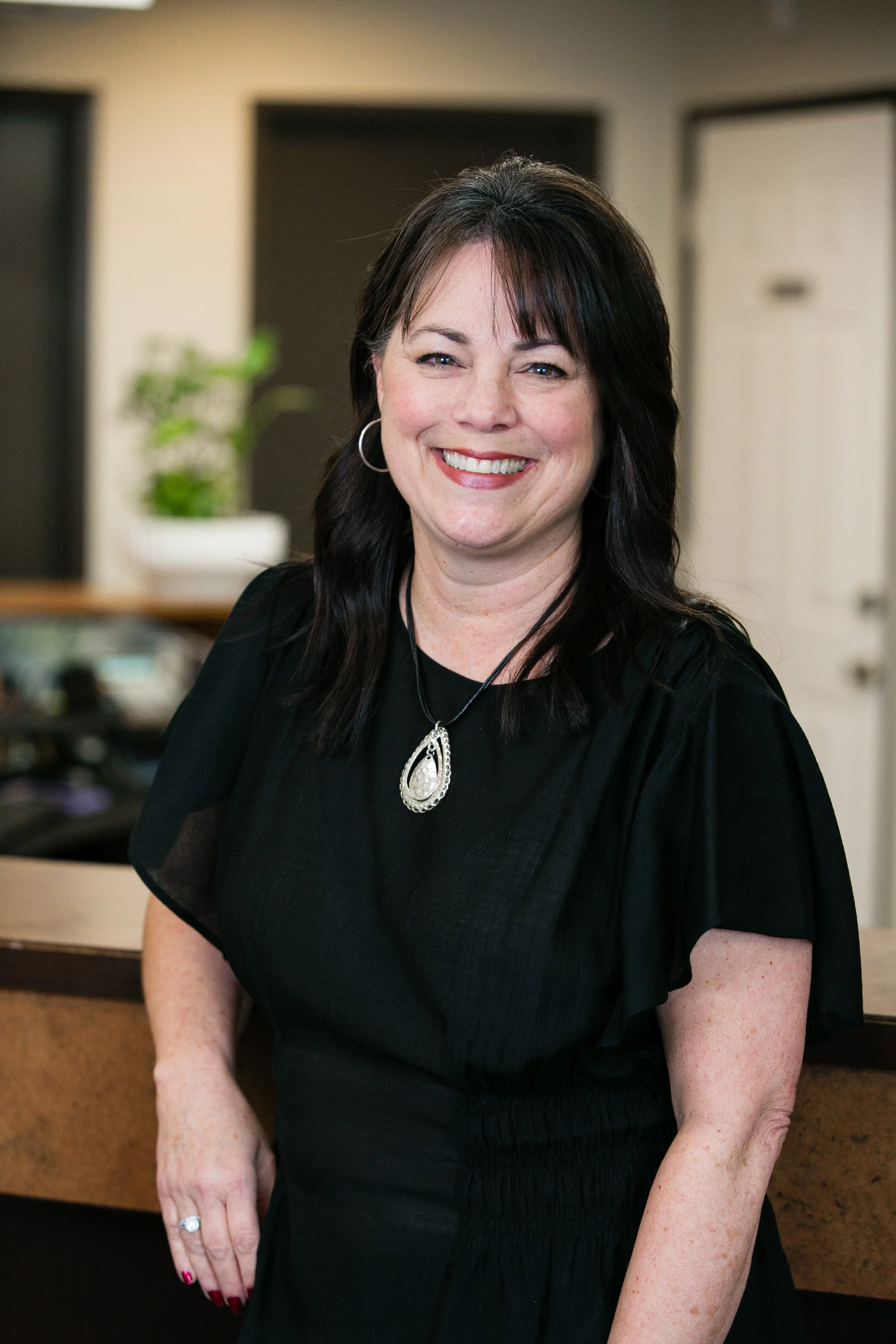 A smiling woman with dark hair and hoop earrings, wearing a black dress and a necklace, standing indoors.