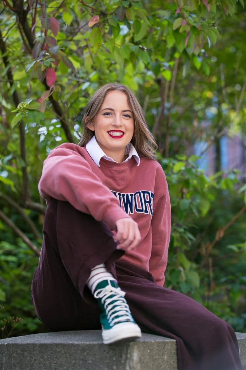 Young woman with shoulder-length brown hair, wearing a maroon sweatshirt with lettering, white collared shirt underneath, maroon pants, and teal sneakers, sitting outdoors on a concrete surface surrounded by green foliage, smiling at the camera.