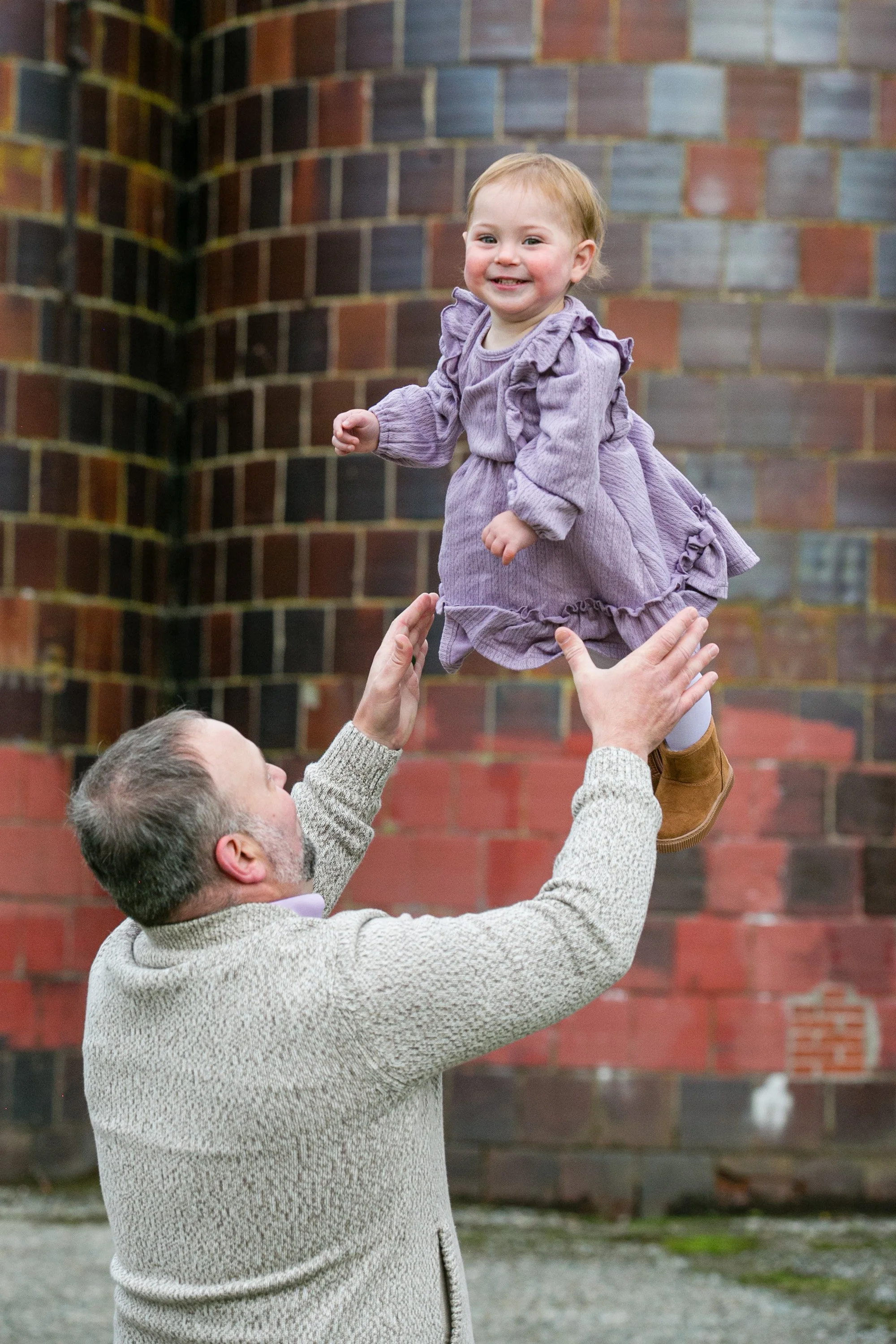 A man lifting a young girl in the air outdoors with a brick wall background.